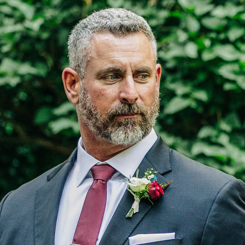 A man with gray hair and beard, wearing a dark suit, white shirt, and red tie, with a boutonnière of red and white flowers, looking to the side outdoors with green foliage in the background.