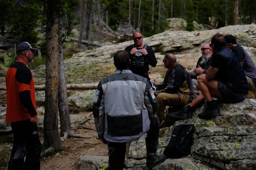 A group of people gathered outdoors in a forested area, sitting on rocks and standing, engaged in conversation.