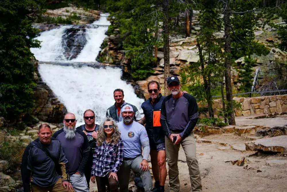 Group of seven people on a rocky trail with a waterfall in the background surrounded by trees.