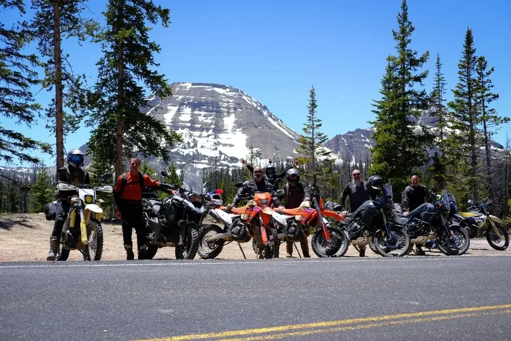 Group of motorcyclists with their bikes parked on the side of a road, with snow-capped mountains and tall pine trees in the background.