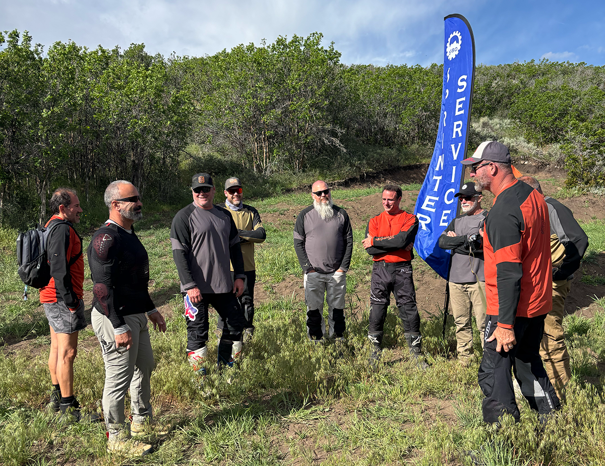 Group of nine men standing outdoors near a blue flag that reads 'SERVICER'. They are dressed in outdoor gear, some wearing motocross or mountain biking attire, and are smiling and talking. The background features green trees and a partly cloudy sky.