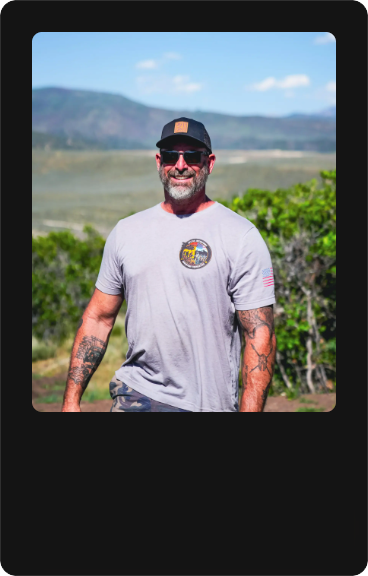 A smiling man with sunglasses, a beard, and tattoos on his arms, standing outdoors with a landscape of hills and trees in the background.