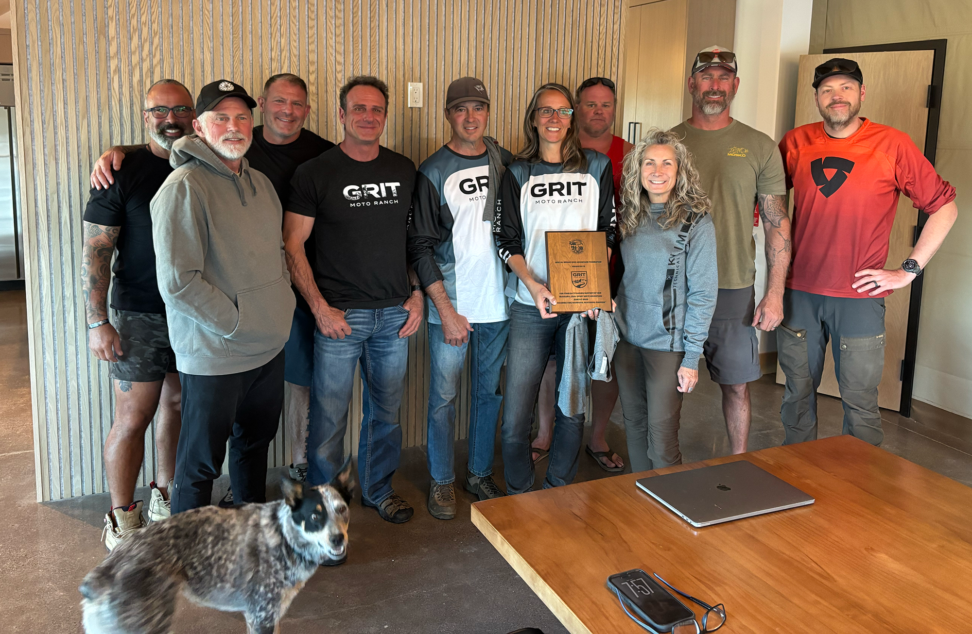Group of eleven people, including men and women, standing together indoors, some wearing casual clothing and others in branded shirts, holding a plaque. A dog is partially visible in the foreground. A wooden wall and a door are in the background; a table with a laptop, phone, and glasses is in the foreground.