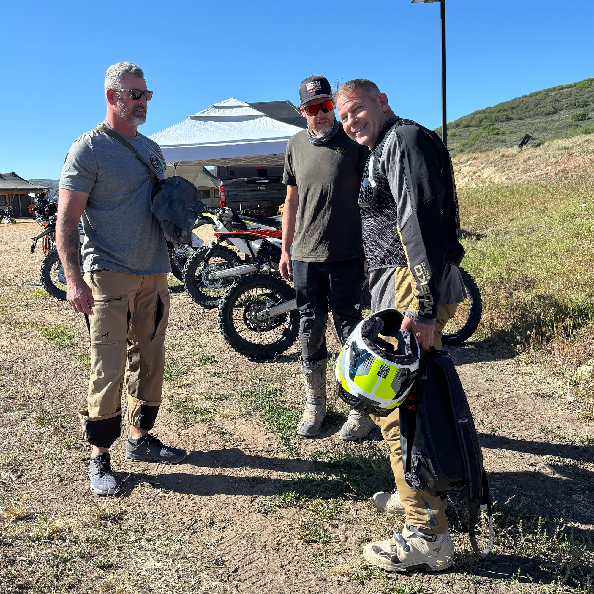 Three men standing outdoors near dirt bikes under a white canopy, with a hillside covered in greenery in the background, on a clear sunny day.