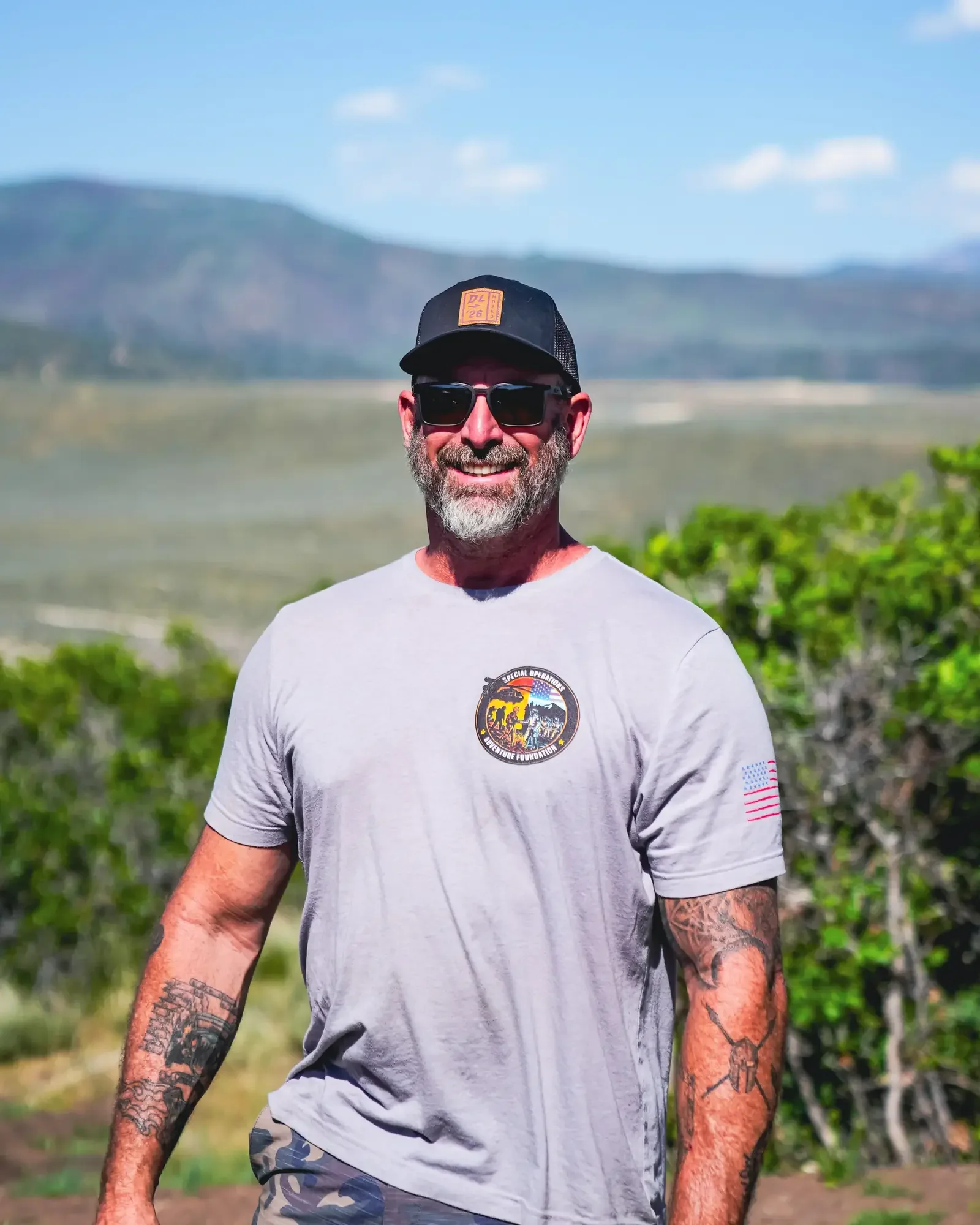 A smiling man with a beard, wearing sunglasses, a gray T-shirt with a badge, camouflage shorts, and a black cap, standing outdoors in a vineyard with mountains in the background under a blue sky.