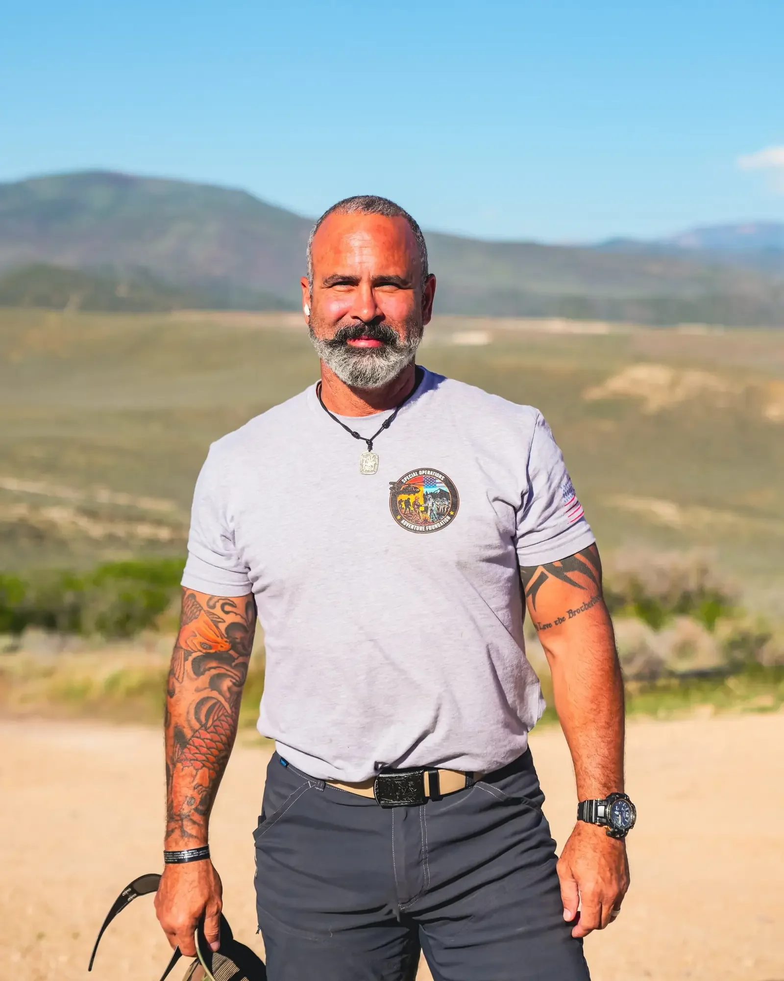 A middle-aged man with a beard and tattoos, standing outdoors with mountains in the background. He is wearing a light gray t-shirt with a patch, dark pants, sunglasses in his right hand, a black wristwatch, and a dog tag necklace, smiling slightly and squinting in the sunlight.