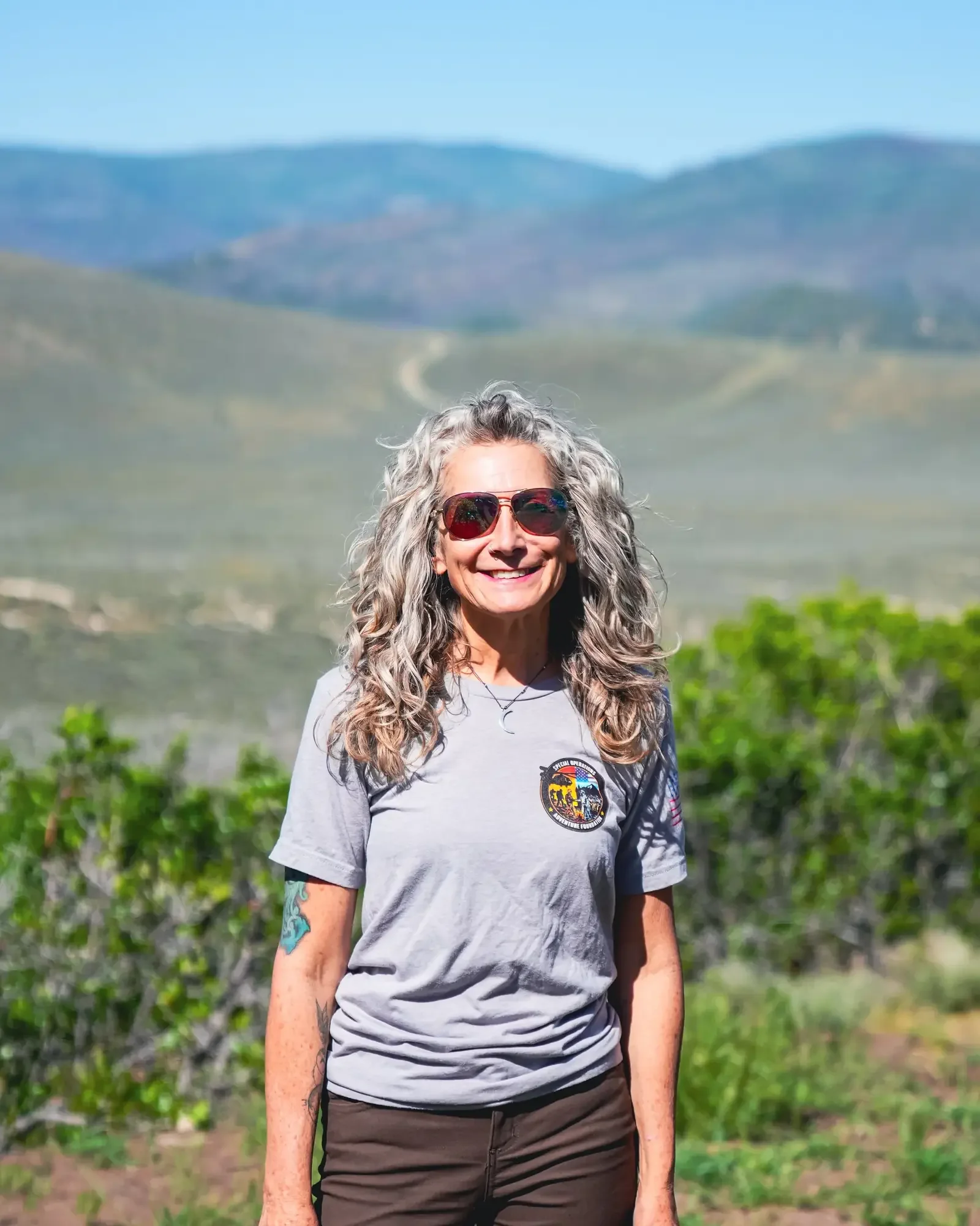 Smiling woman with long, curly gray hair wearing sunglasses, a gray t-shirt, and dark pants outdoors with green hills and mountains in the background.