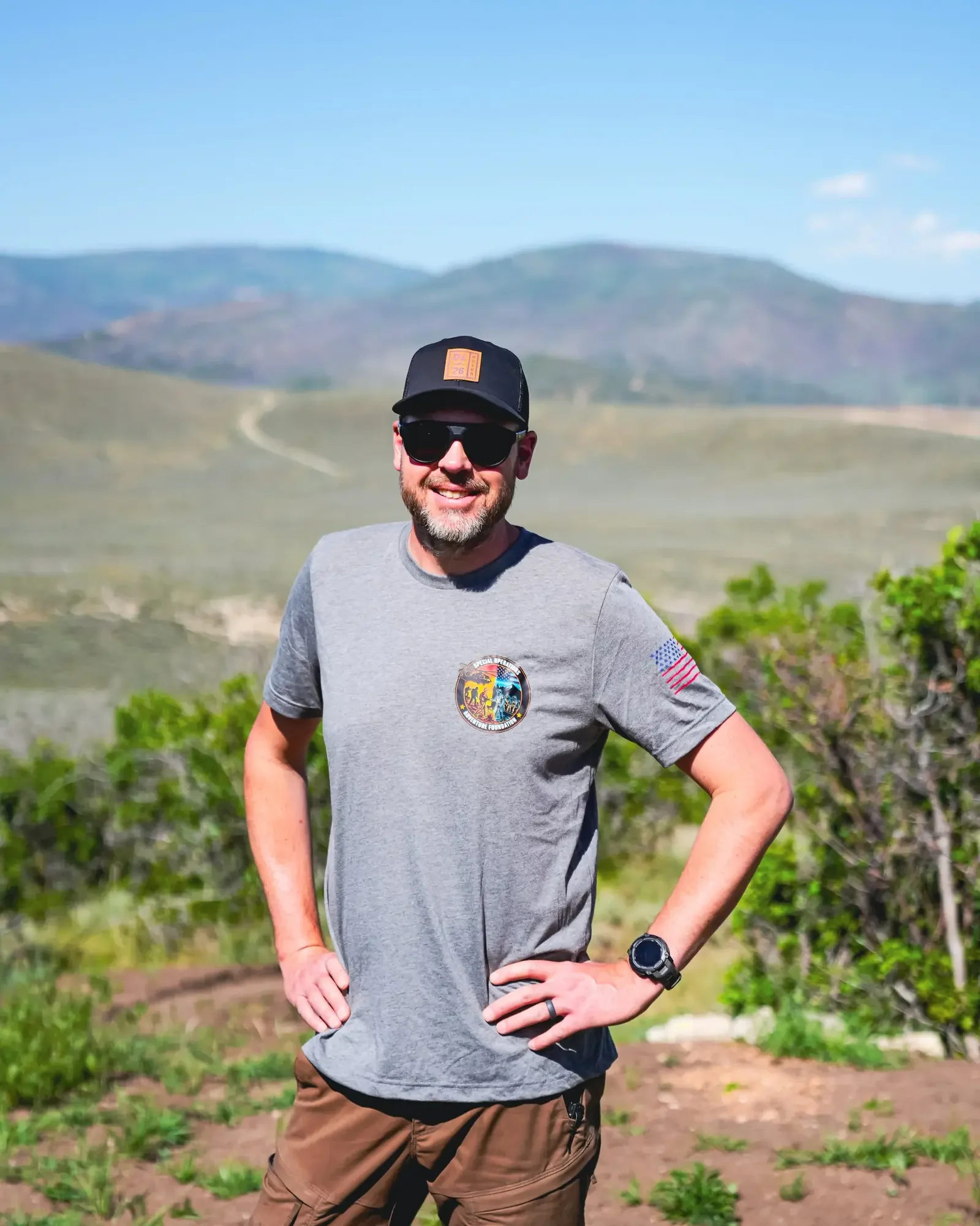 A man standing outdoors in a rural area with hills in the background, wearing sunglasses, a black cap, a grey T-shirt with patches and logos, and brown shorts, smiling with his hands on his hips.