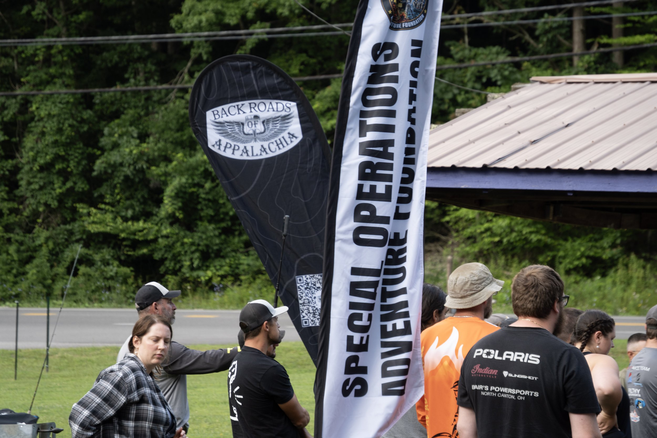 Group of people gathered outdoors near a road with trees in the background, some wearing hats and sports attire. Two vertical banners are displayed, one with the text 'Back Roads of Appalachia' and the other with partially visible text starting with 'RIDE NATION'S'.