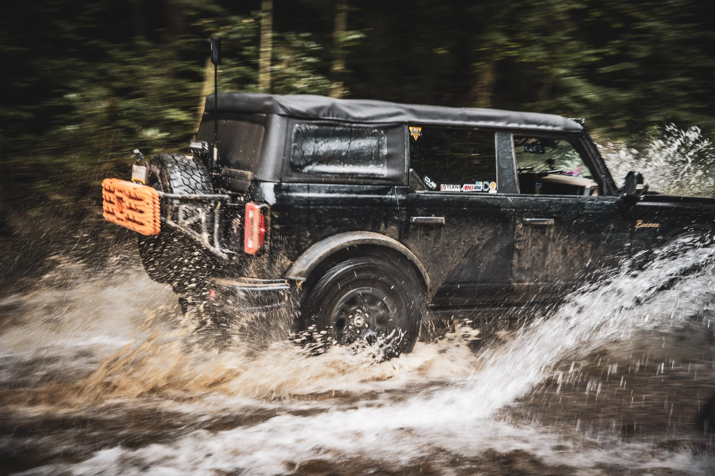A black off-road vehicle driving through a muddy, forested area with water splashing around its wheels.