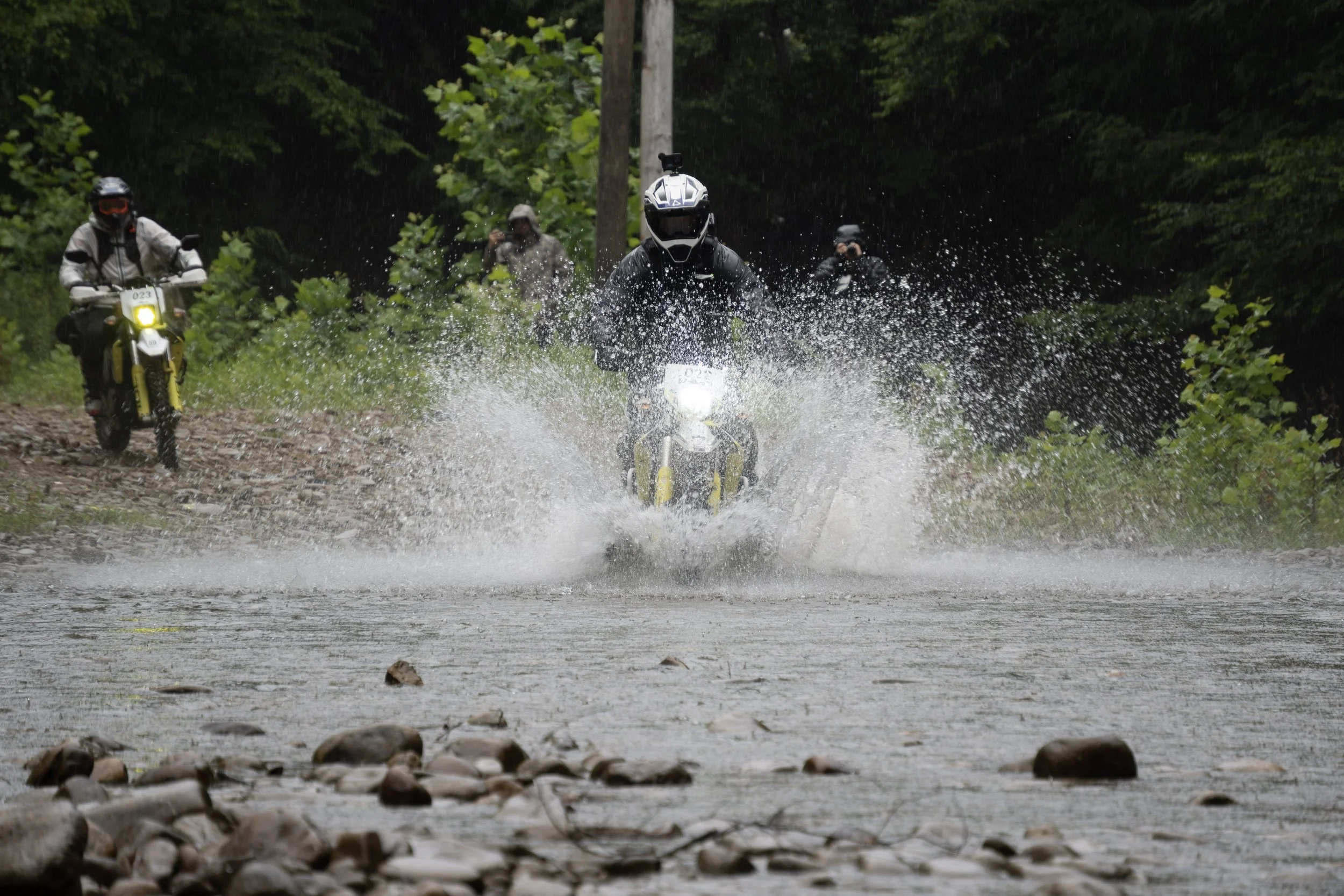 A group of four motorcyclists riding motorcycles through a shallow stream in a dense forest. The central rider is splashing water as they ride through the water, with two other riders visible in the background and one on the left side of the image.