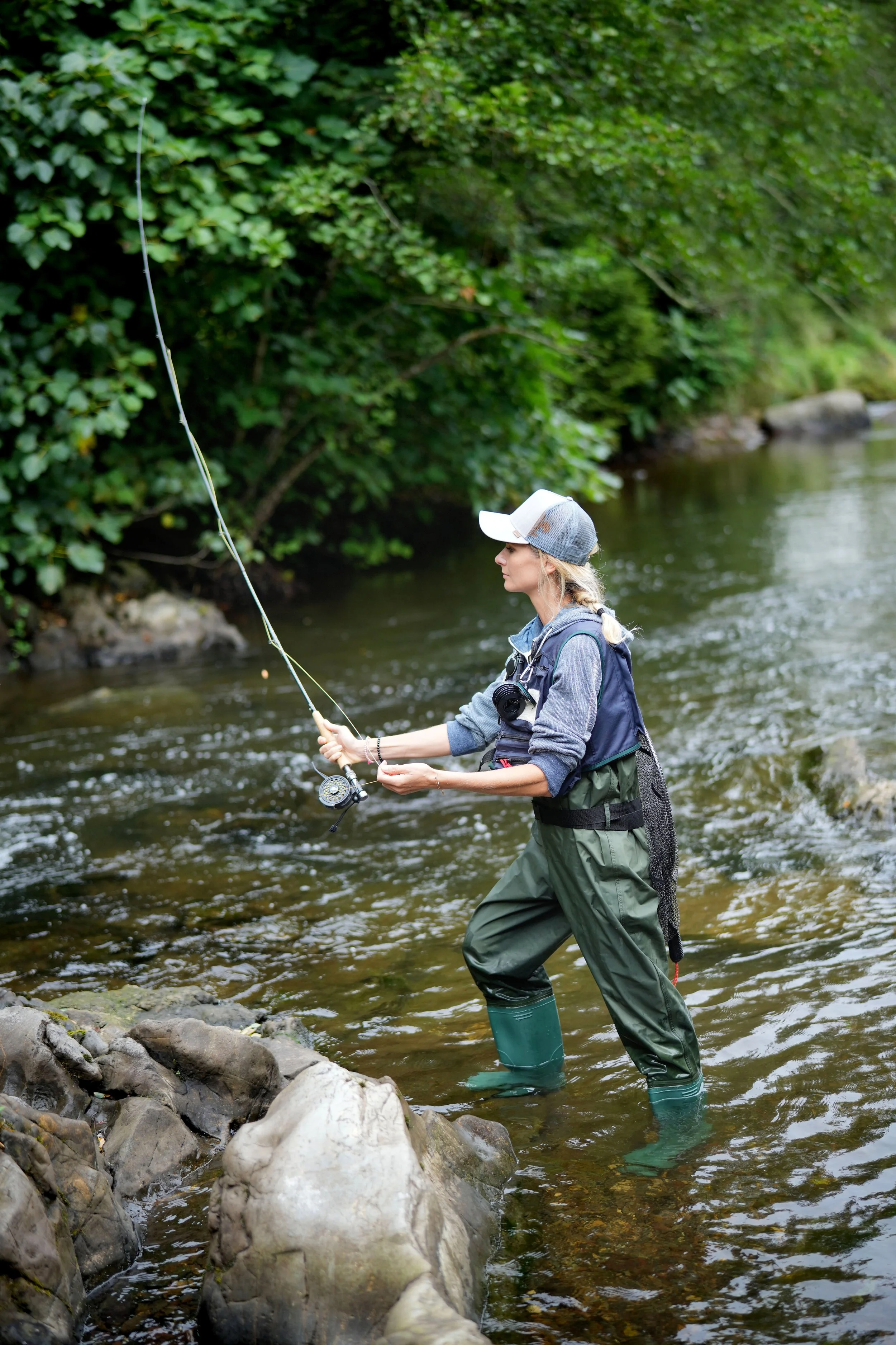 Woman fishing in a river surrounded by lush green trees, wearing a cap and waterproof waders.