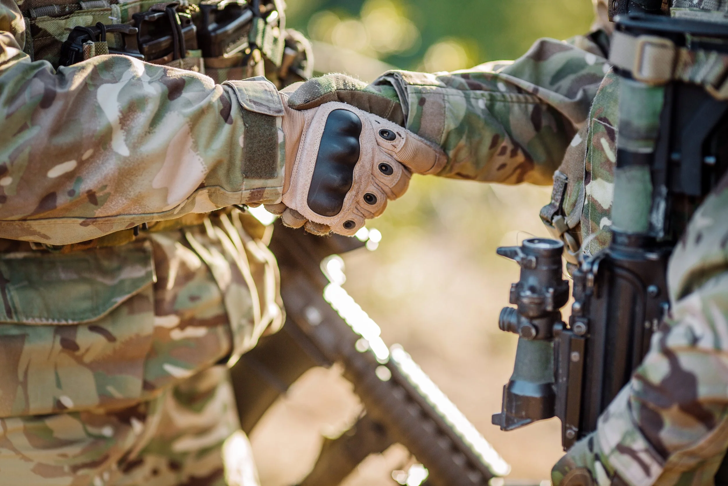 Two soldiers in camouflage uniforms shaking hands outdoors, with one soldier wearing tactical gloves and equipment, and a firearm is visible attached to the uniform.