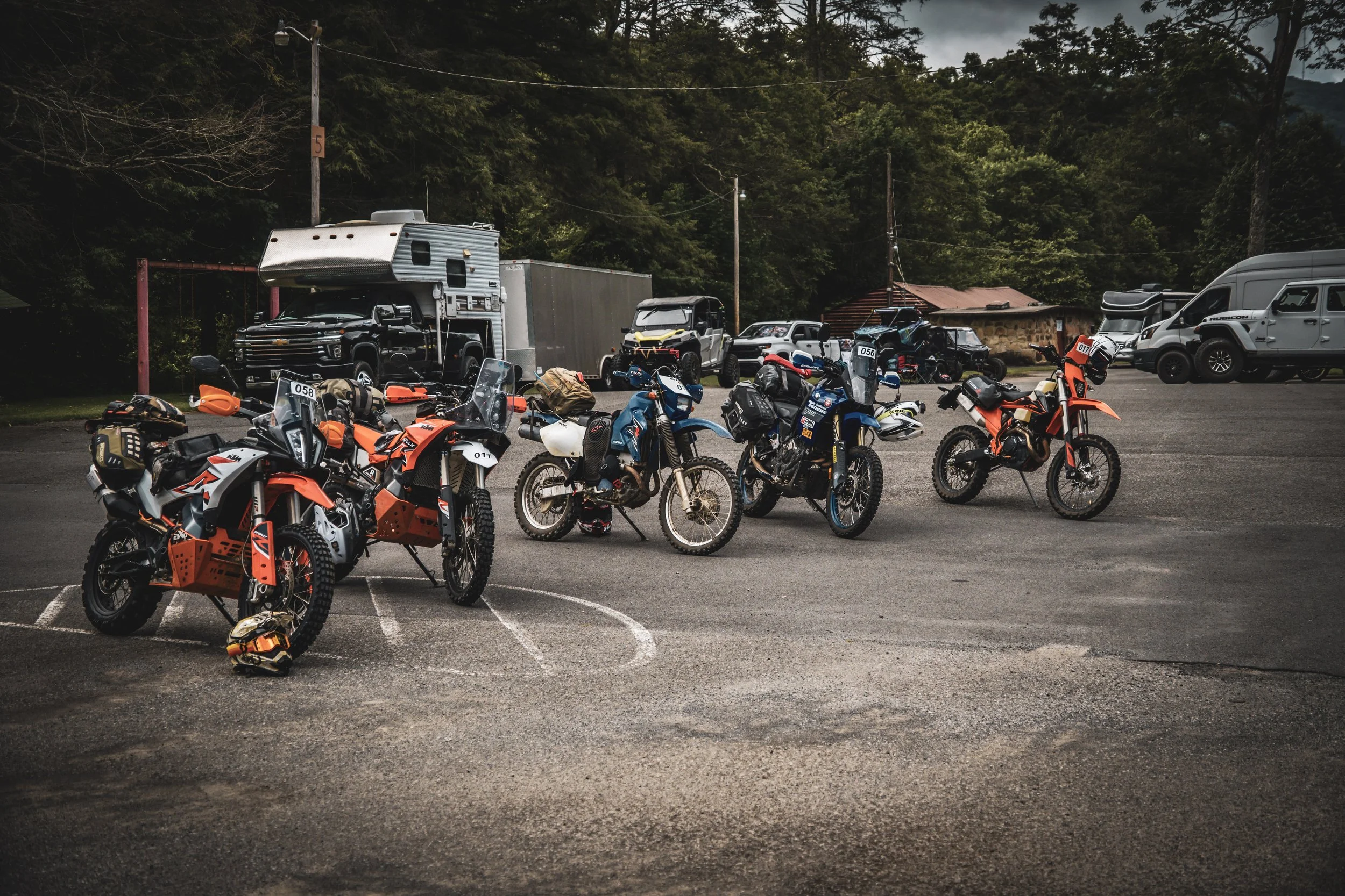 A parking lot filled with off-road motorcycles and nearby vehicles, with a wooded hillside in the background.