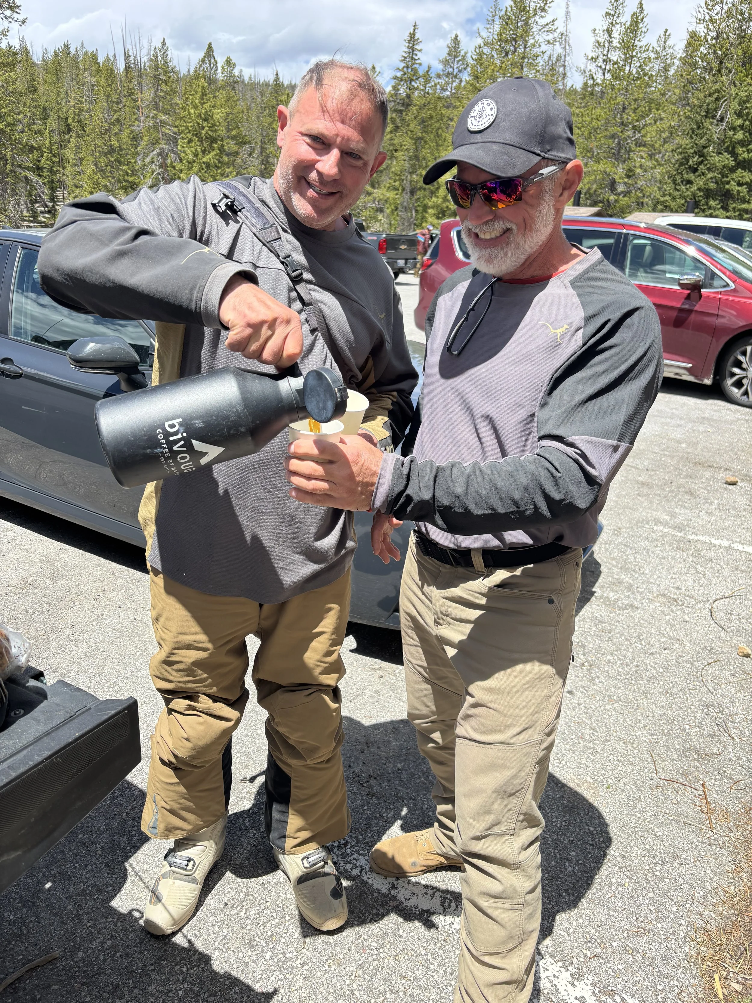 Two smiling men outdoors, one pouring coffee into a cup and the other holding it, with cars and green trees in the background.