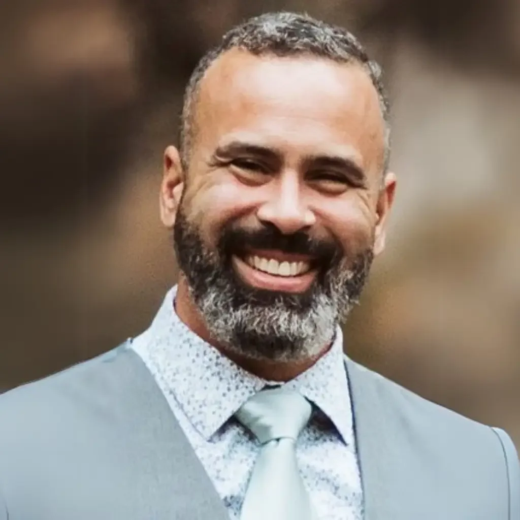 A man with a beard and mustache smiling, dressed in a suit with a shirt and tie, standing outdoors.
