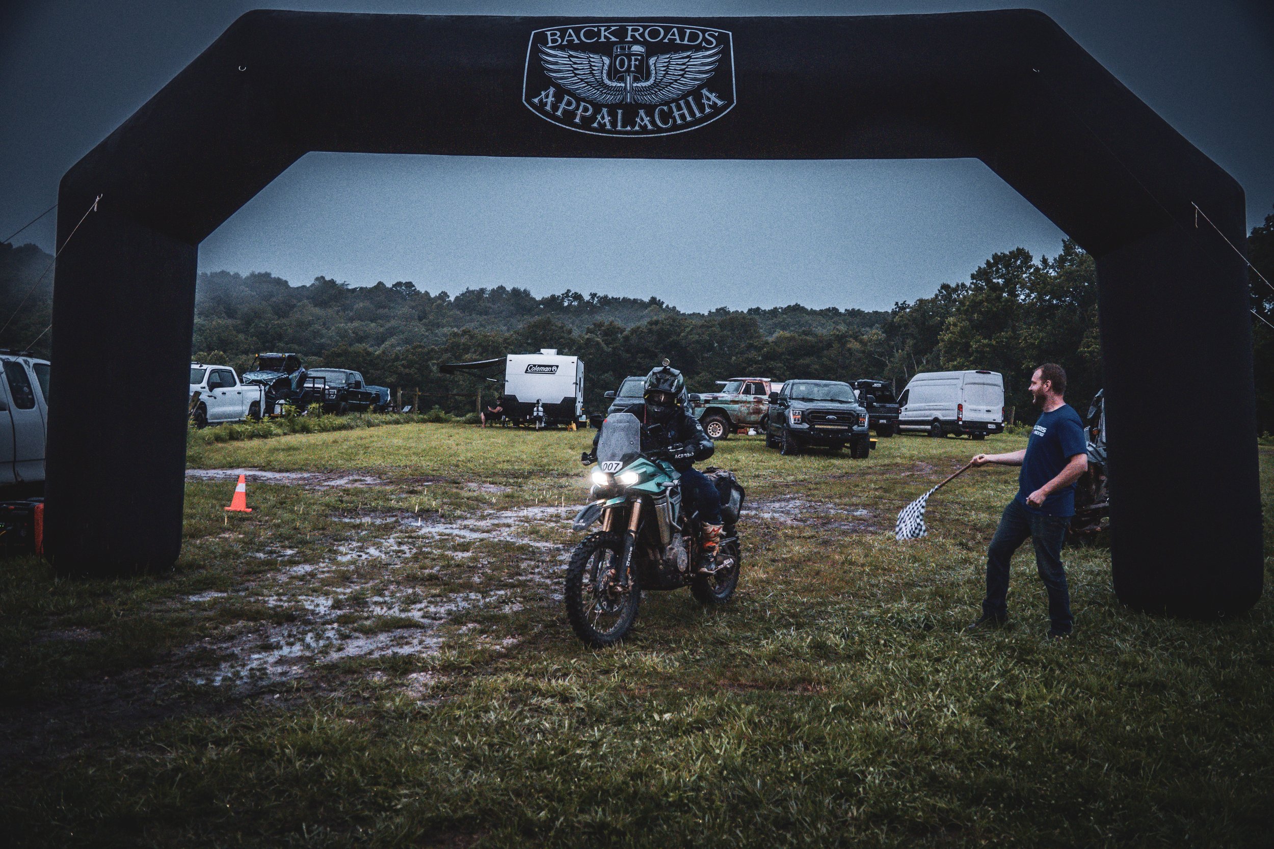 An outdoor event space with a large inflatable arch reading 'Back Roads of Appalachia' in black with white text and design. Behind the arch, there are several parked vehicles, including trucks, SUVs, and a camper trailer, on a grassy area under a cloudy sky. In the foreground, a person on a motorcycle and a person in a blue shirt holding a checkered flag are visible, and there are orange traffic cones on the muddy ground.