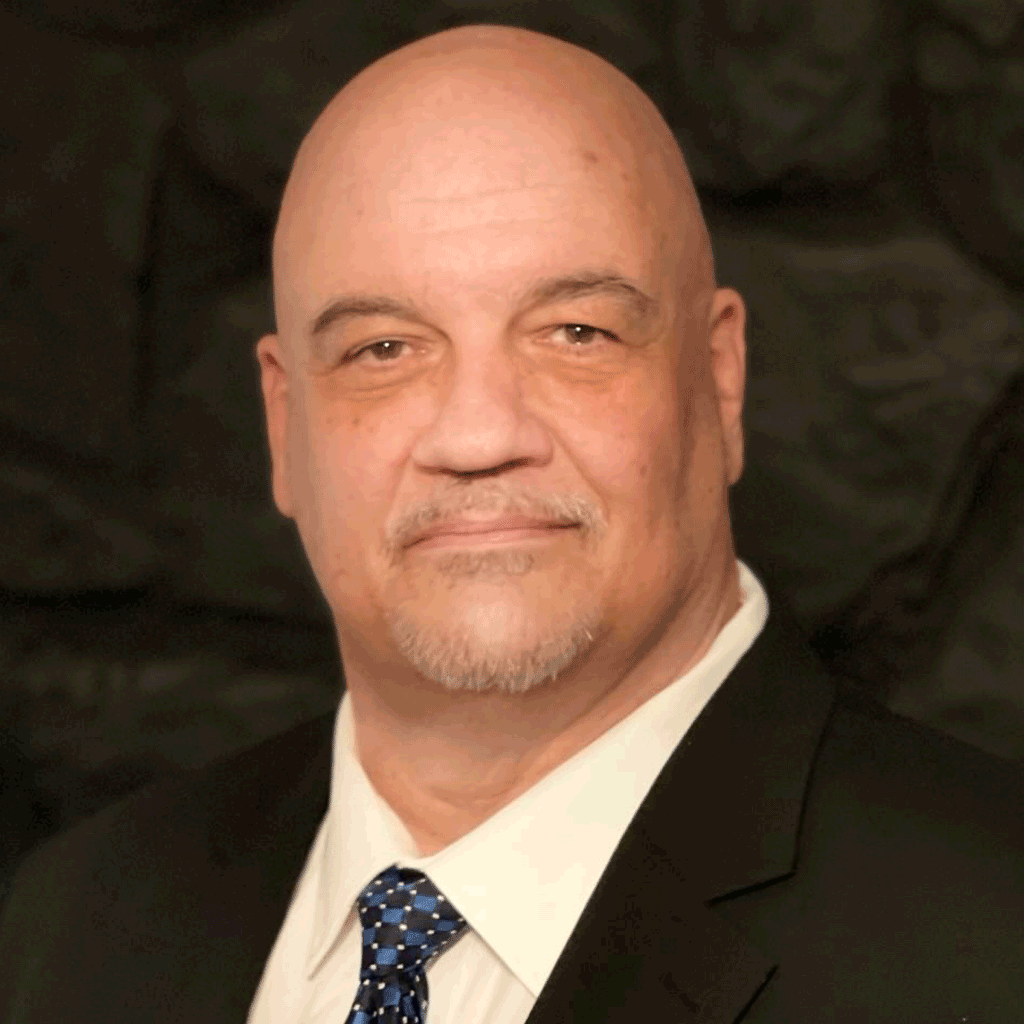 Close-up portrait of a middle-aged man with a bald head, light facial hair, wearing a black suit, white shirt, and a blue patterned tie, against a dark background.