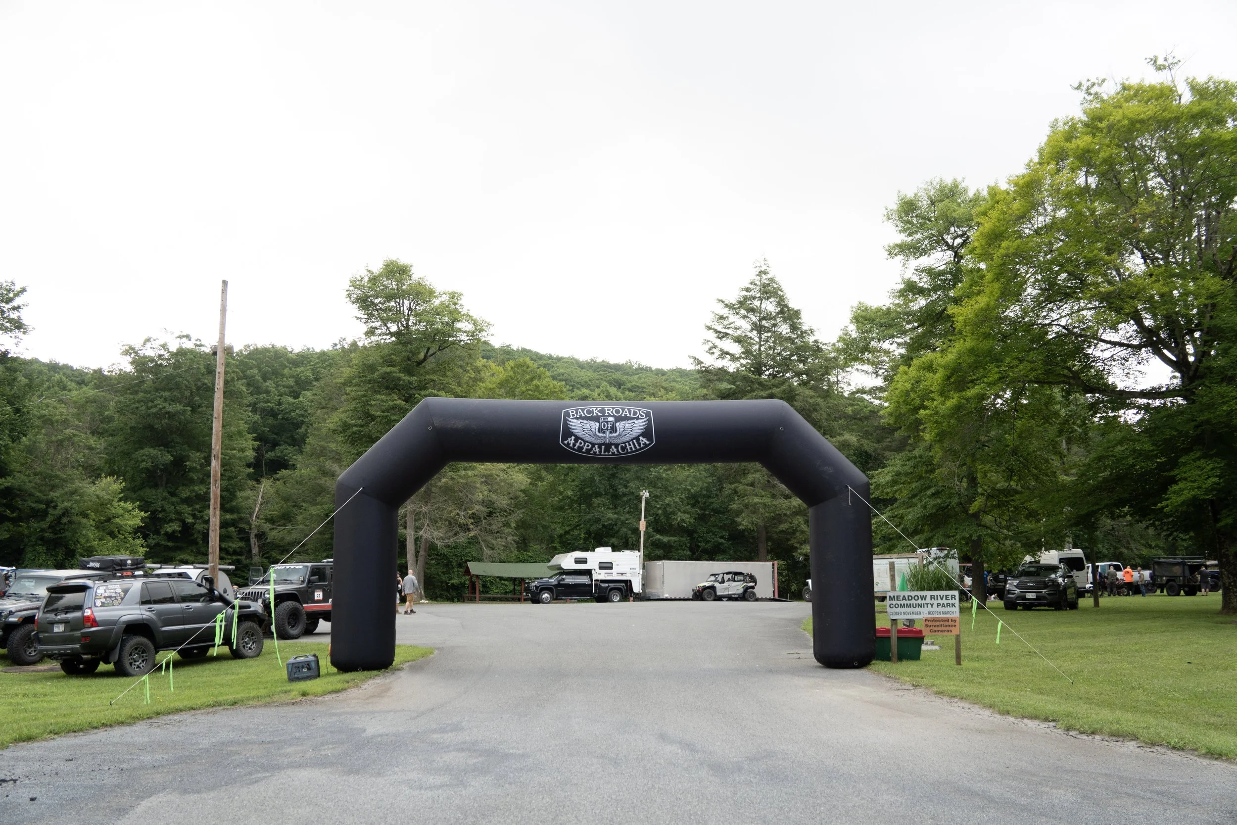 An outdoor scene at Meadow River Community Park with an inlet archway that reads 'Back Roads of Appalachia.' There are numerous parked cars and RVs, a volleyball net, and green trees in the background.