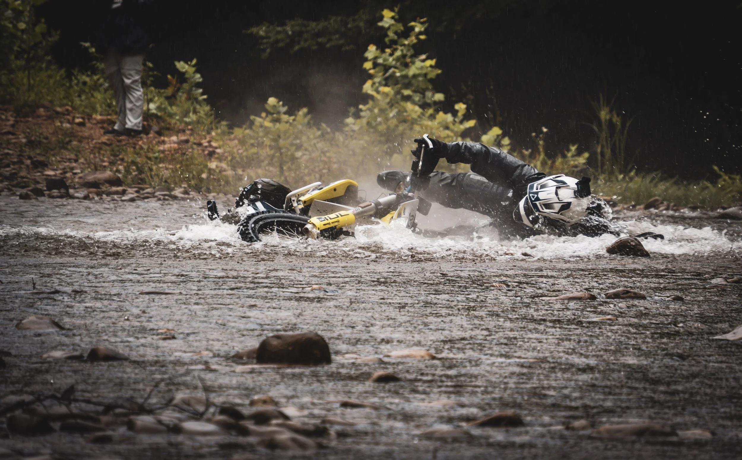 A person dressed in black riding a yellow and black off-road motorcycle crashes into a river, falling into the water on a rainy day, with trees and water surrounding the scene.