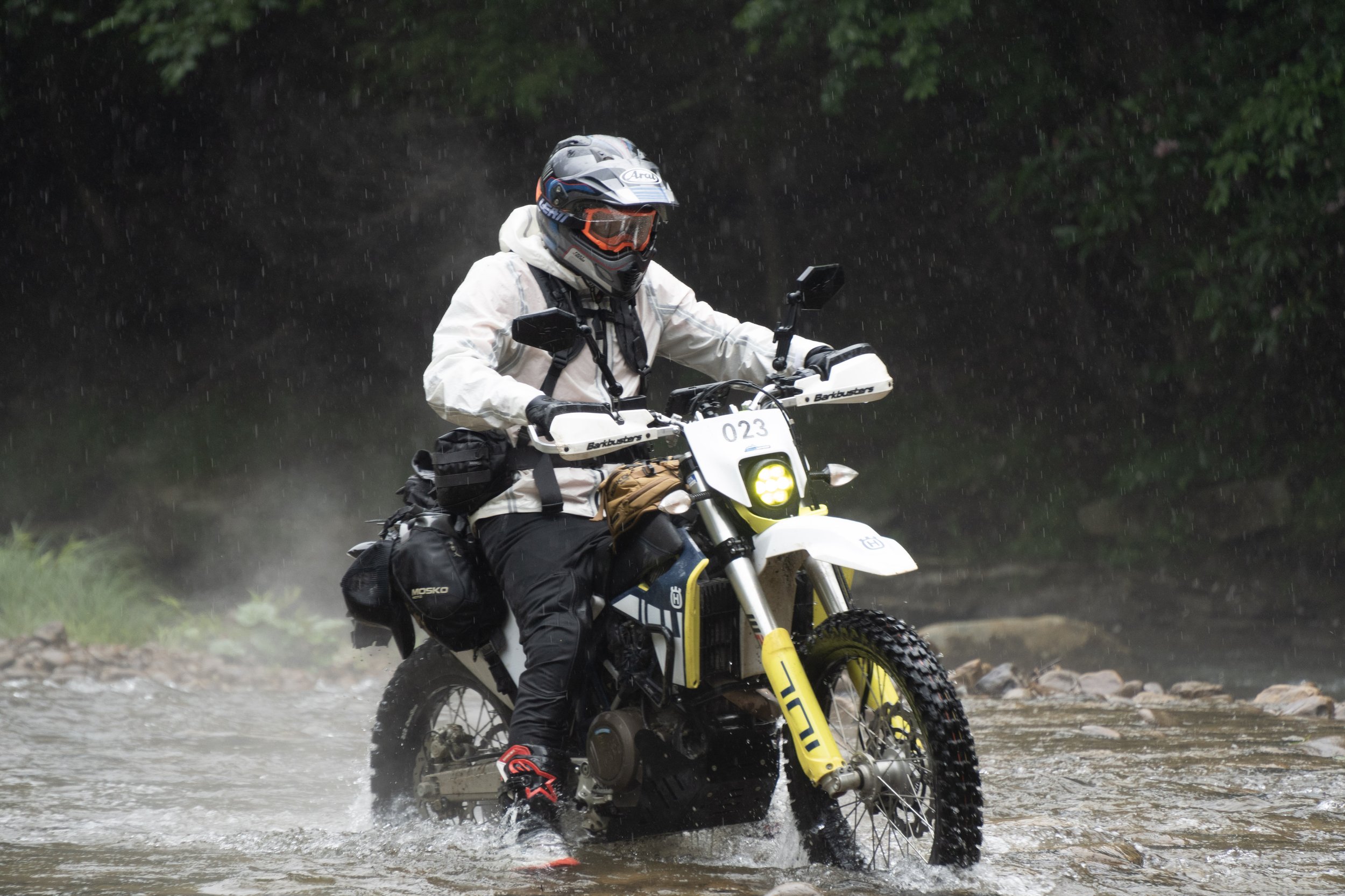 Motorcycle rider wearing protective gear and helmet crossing a stream during rain.