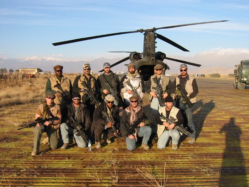 A group of ten armed men in tactical gear posing in front of a military helicopter in an open field with mountains in the background.