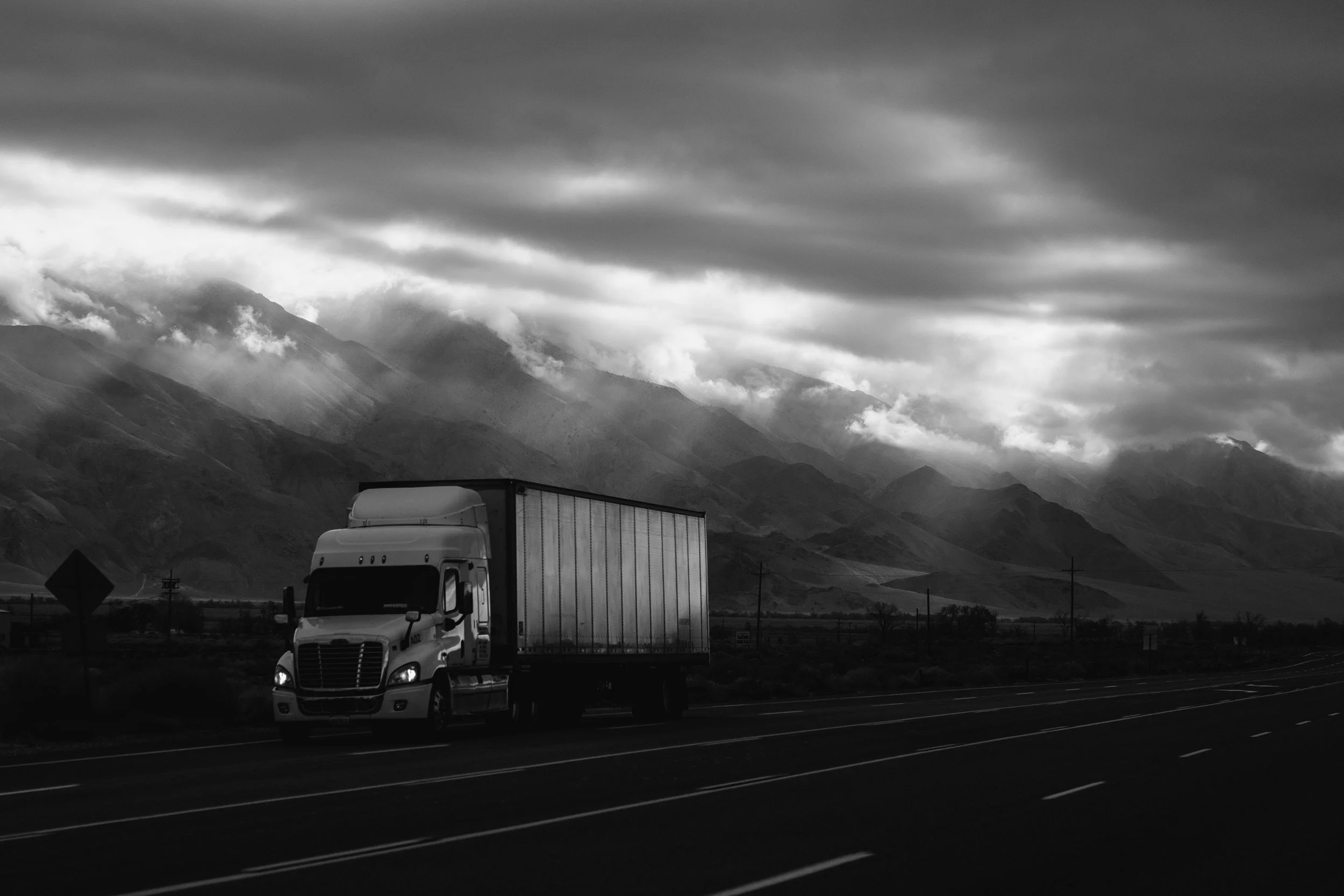 A semi-truck driving on a highway with mountains and cloudy sky in the background.