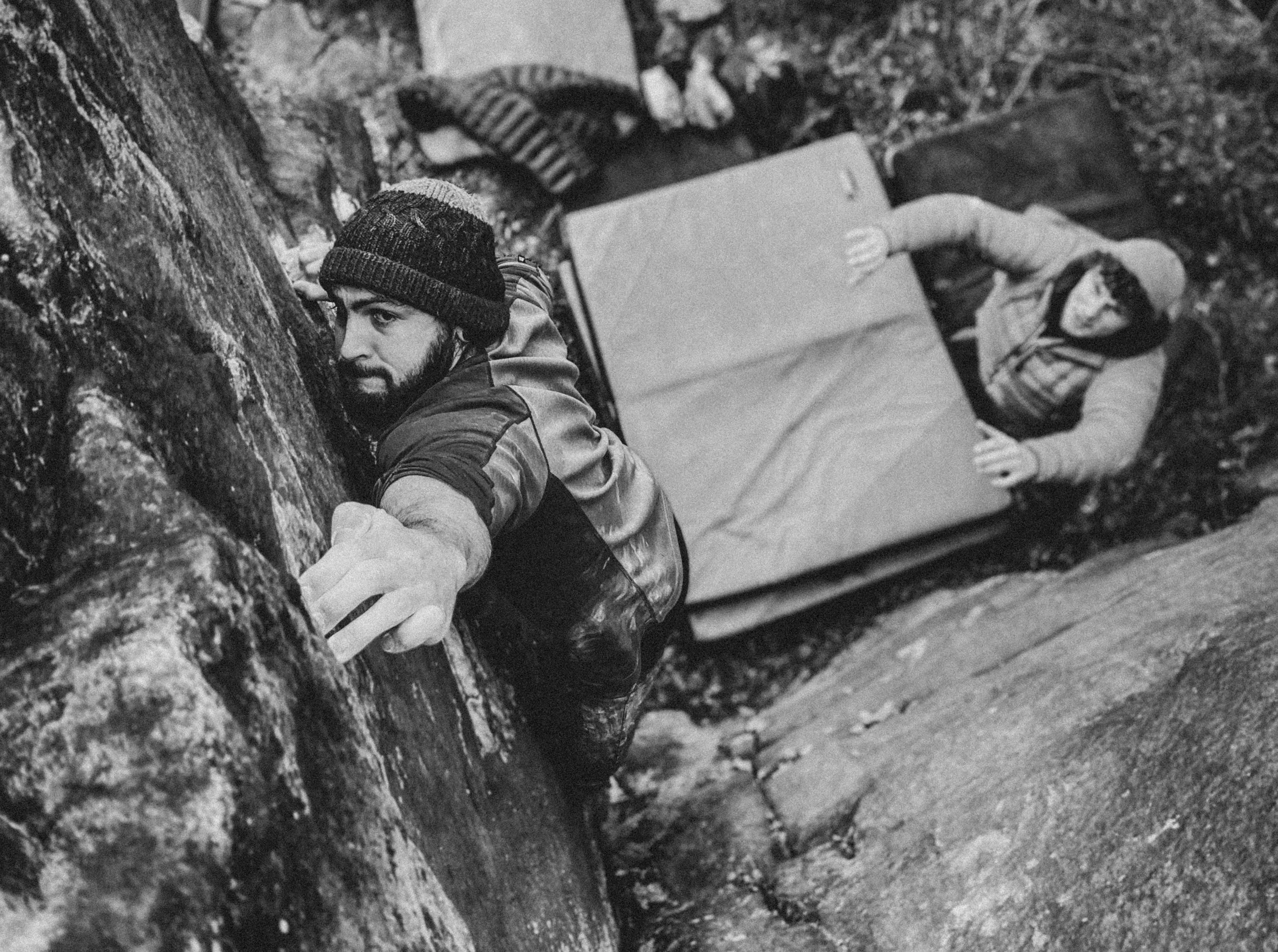 A man with a beard and wearing a knit cap is rock climbing on a textured surface. He is reaching with his right hand while looking sideways. Behind him, another person is lying on a crash pad, looking toward the camera, wearing a jacket and a hoodie.
