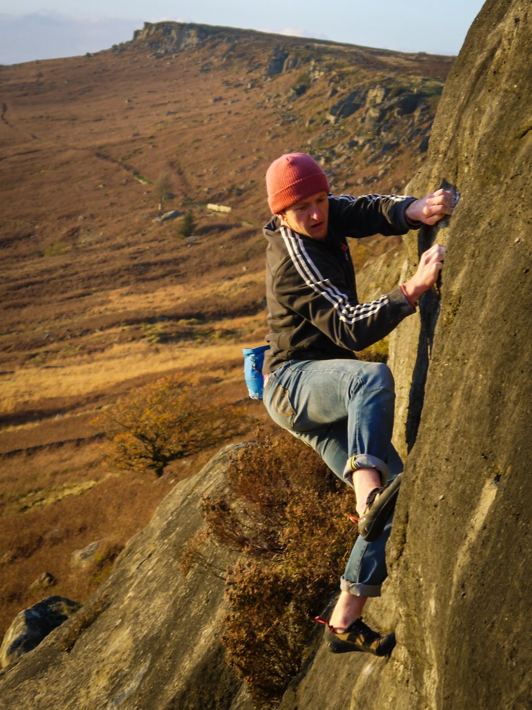 A man rock climbing on a steep outdoor boulder with a mountainous landscape in the background during sunset.