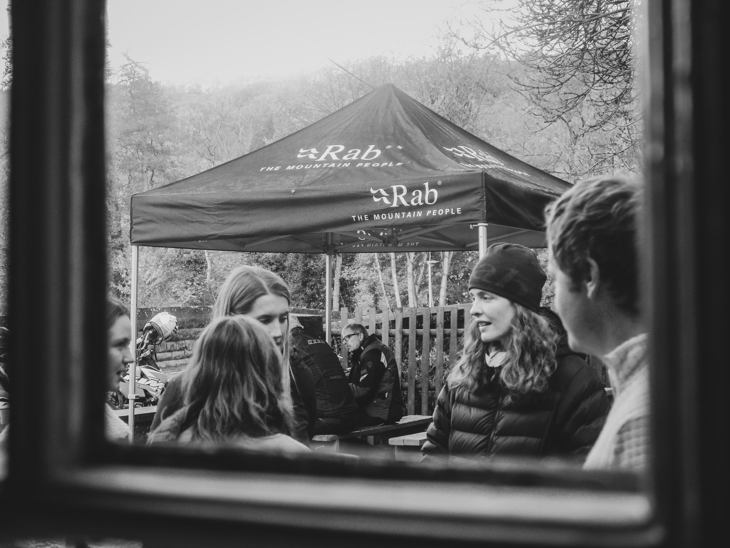 View of people talking outdoors, seen through a window, with a gazebo and trees in the background.