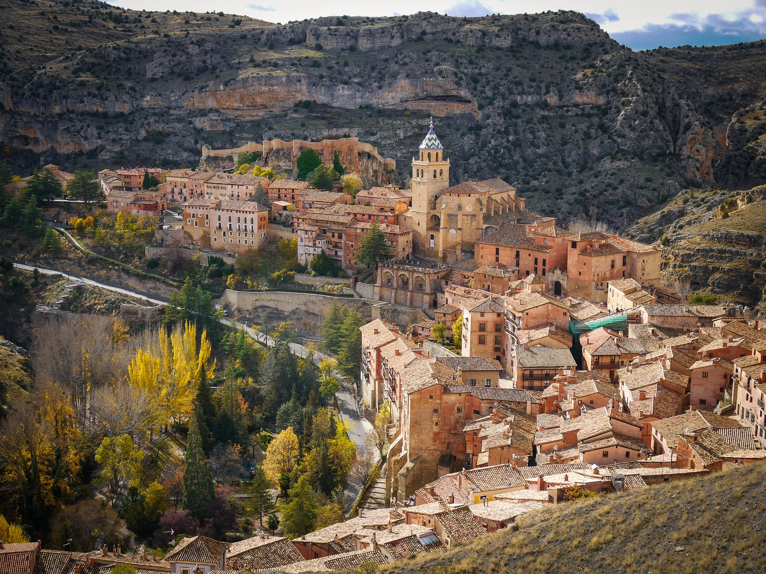 A panoramic view of a historic hillside town with terracotta rooftops, a prominent church with a bell tower, surrounded by rocky hills and lush greenery.