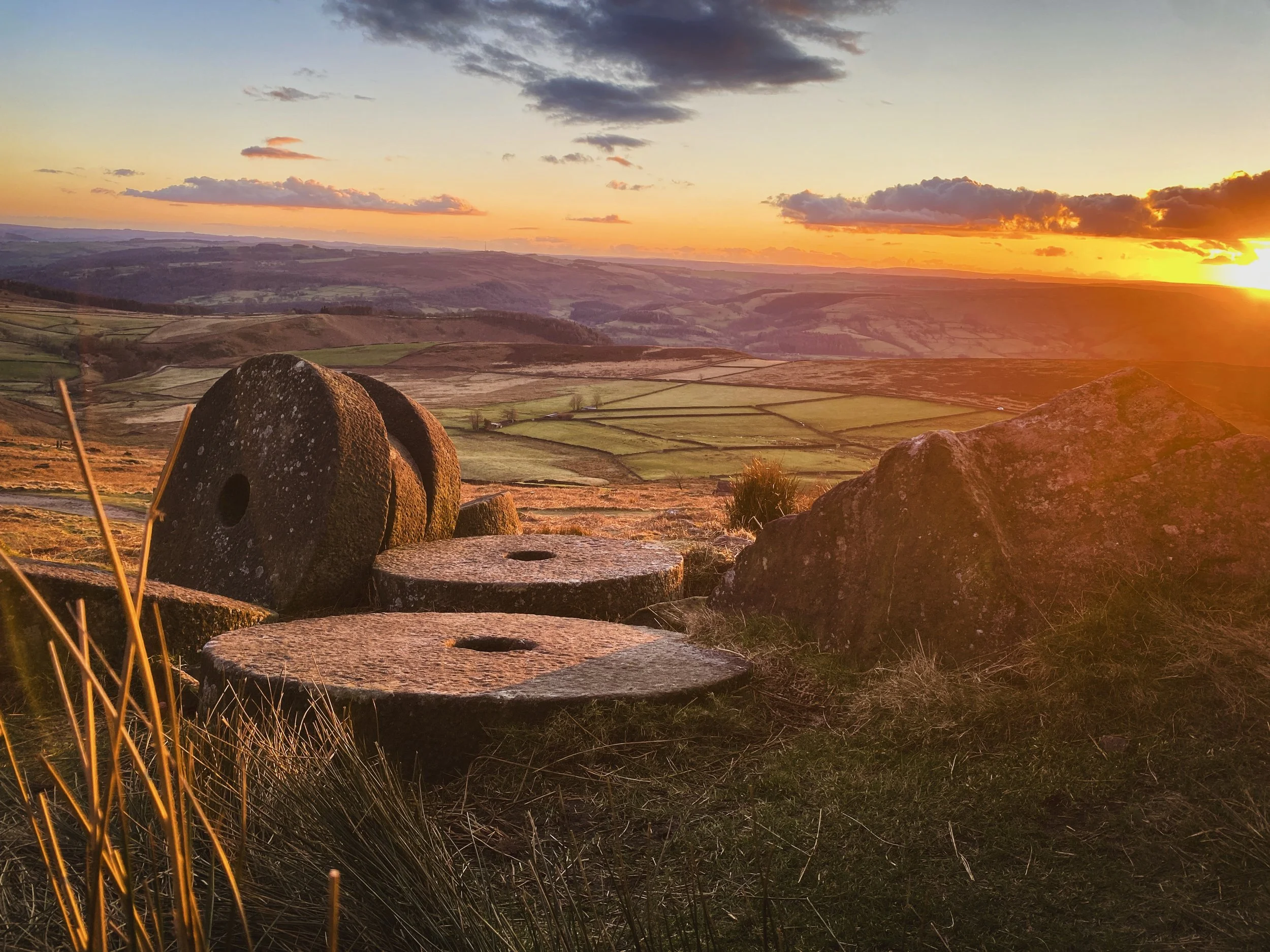 A scenic landscape during sunset with rolling hills, a vibrant orange sky, and large ancient stone circles in the foreground.