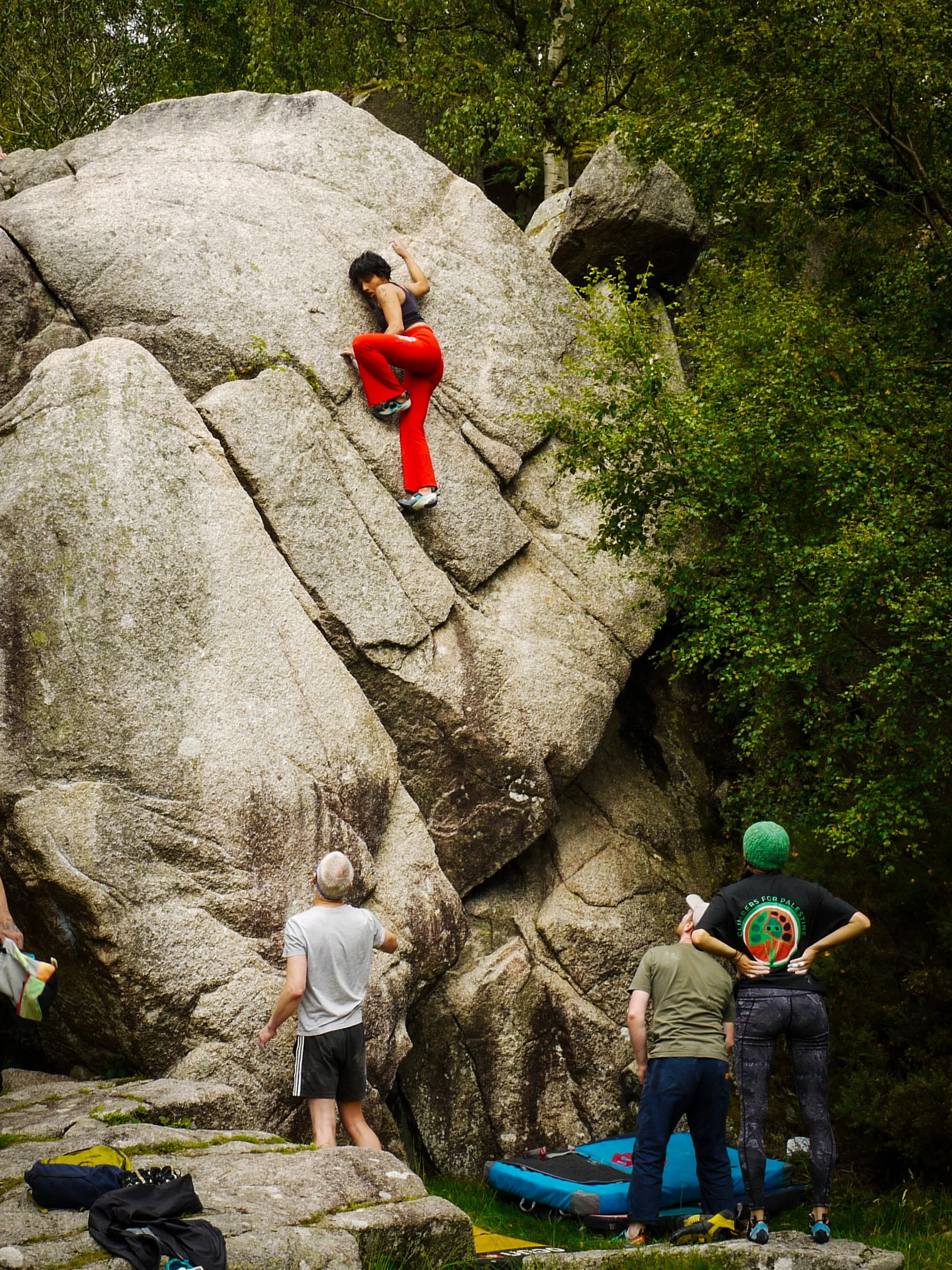A woman climbing a large boulder outdoors, while three people watch nearby. The climber is wearing red pants and a gray top. The scene is surrounded by trees.