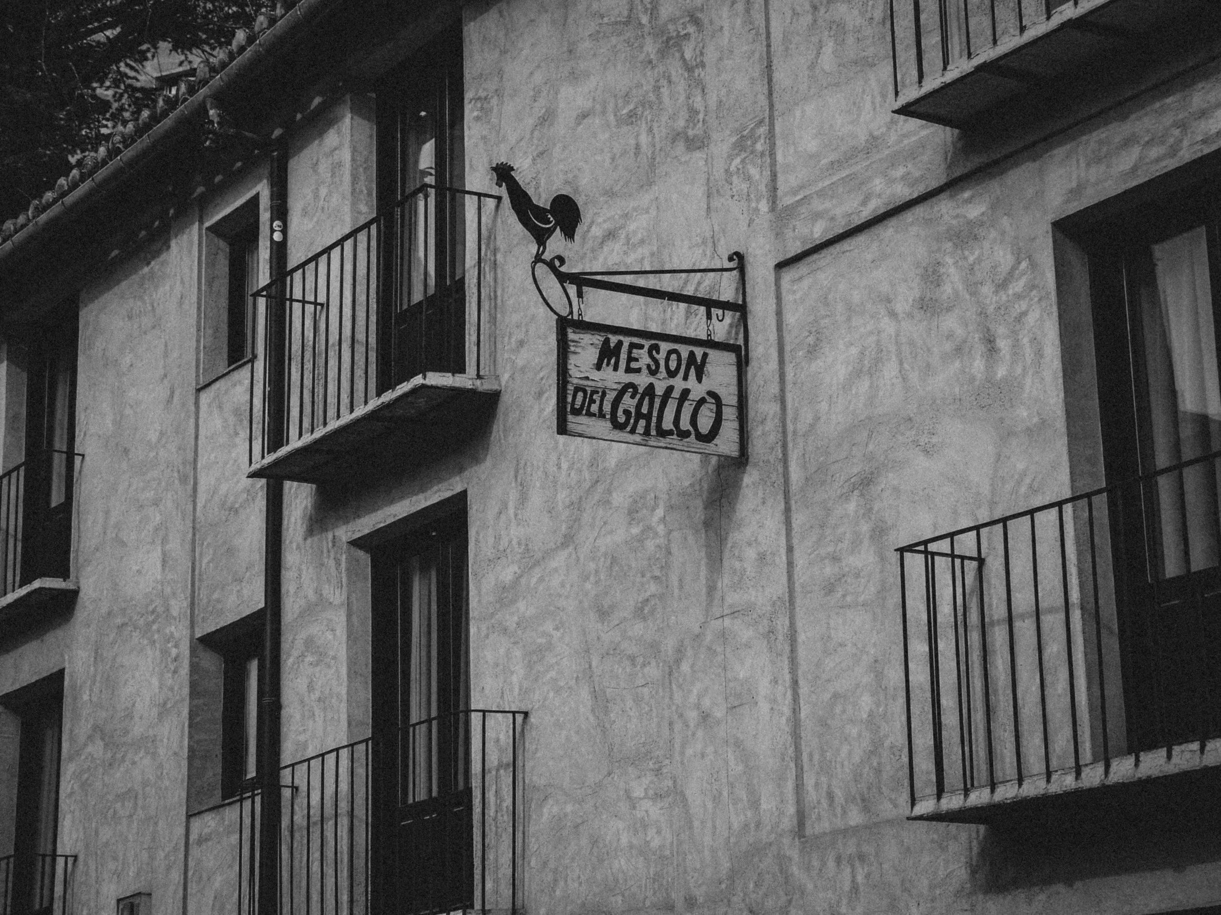 Black and white photo of a building exterior with balconies and a hanging sign that reads 'Meson del Gallo' with a rooster silhouette on top.