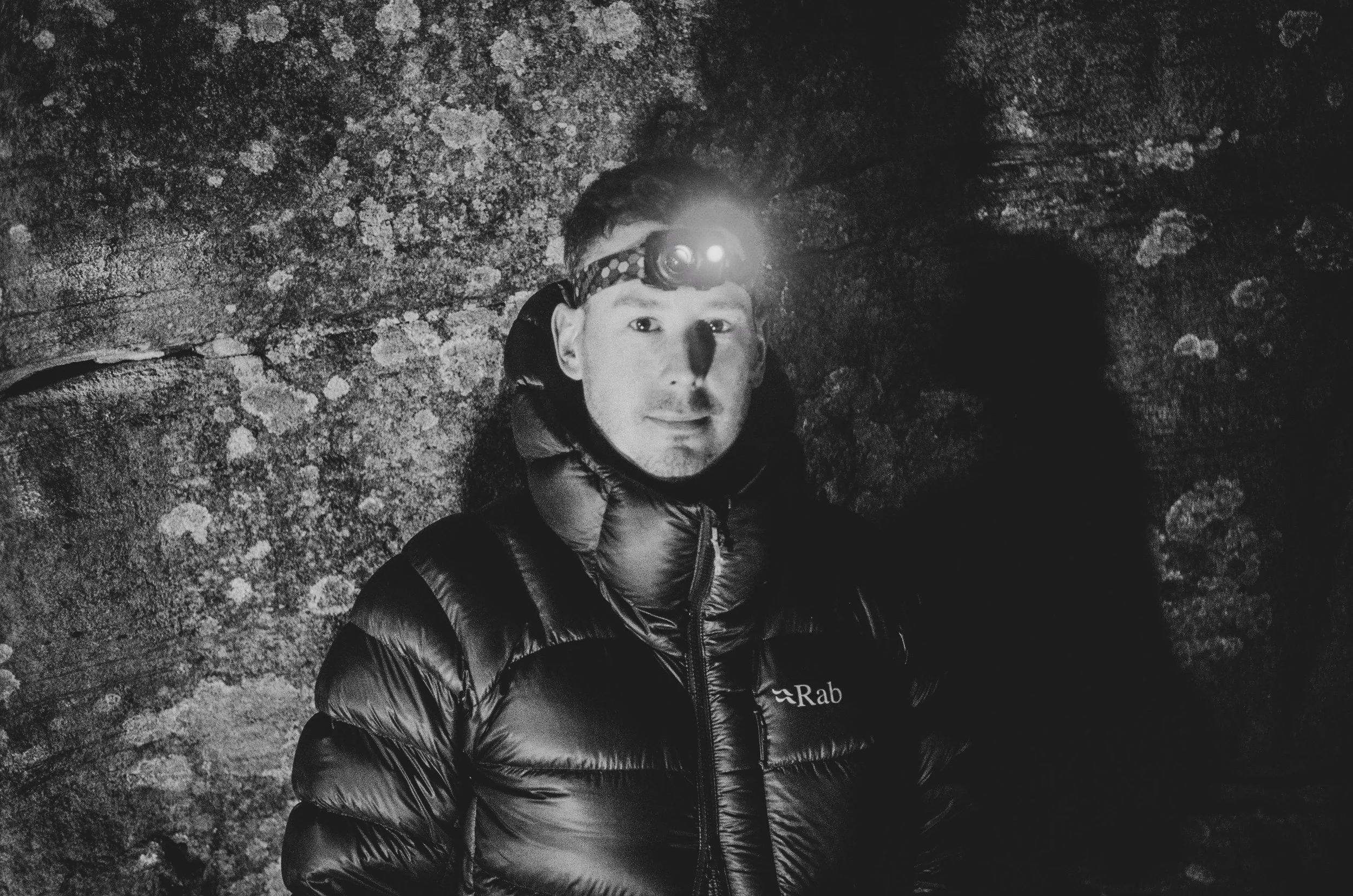 A person wearing a headlamp and a puffy jacket, standing in front of a rocky background in a dark environment.