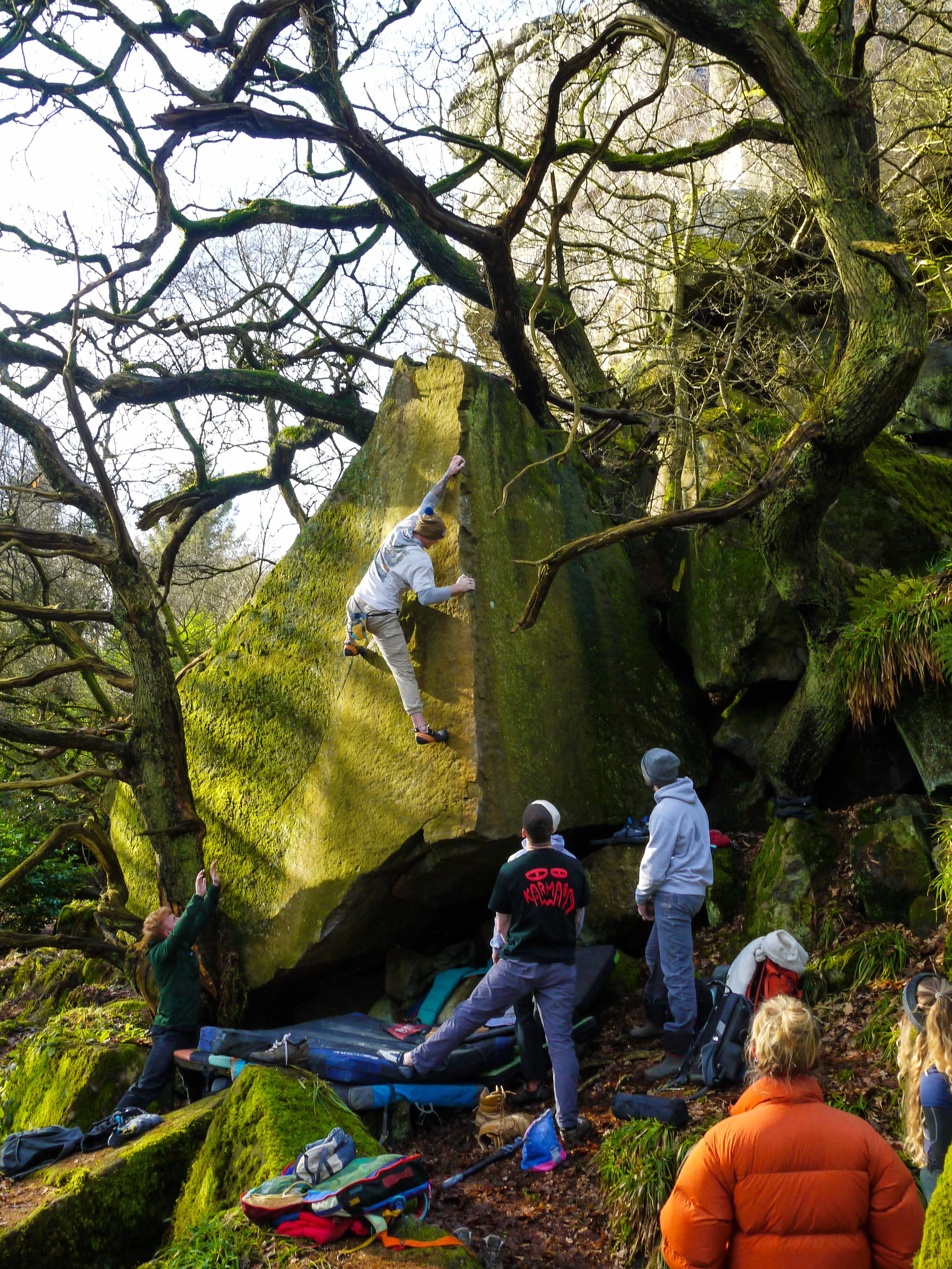 A person rock climbing on a moss-covered boulder in a forest, with several people watching and spotters below.
