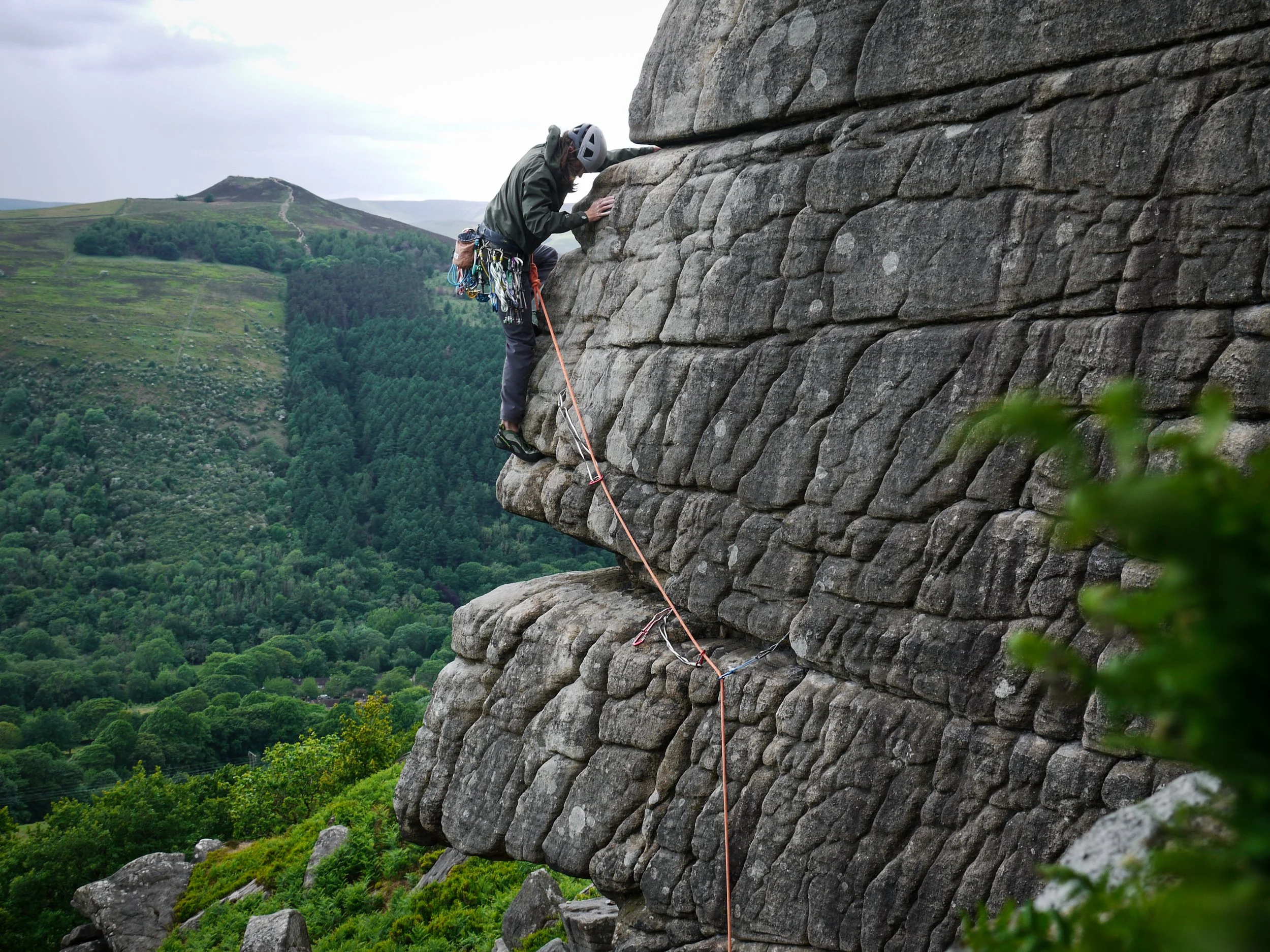 A rock climber ascending an overhanging rock face with a lush green valley in the background.