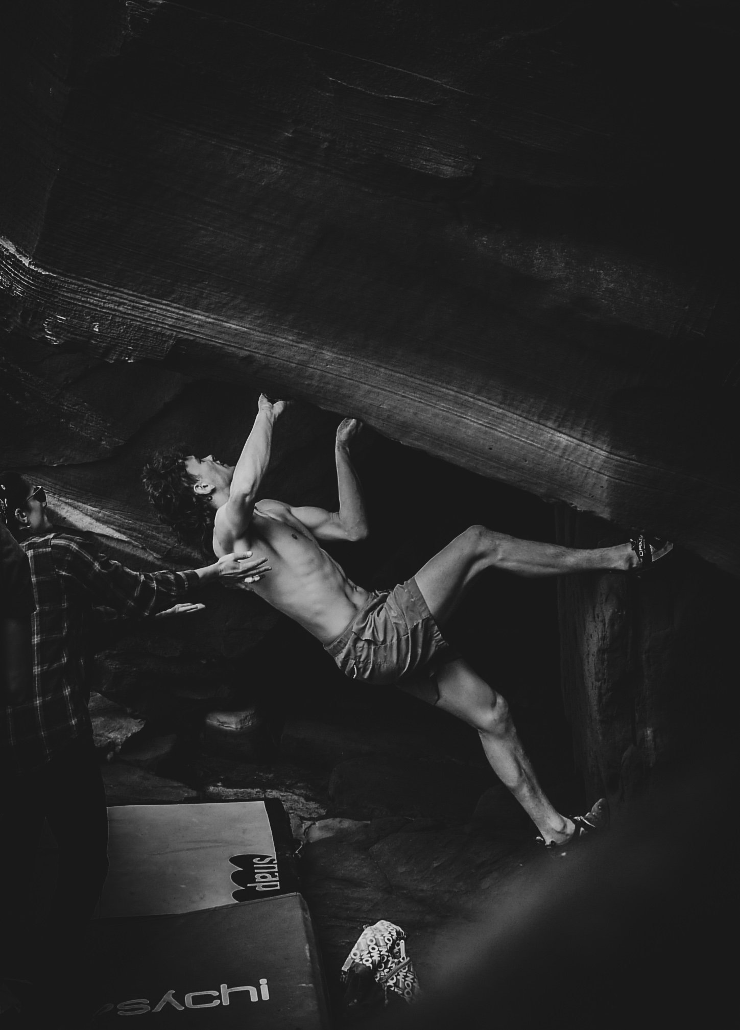 A shirtless man in shorts and climbing shoes is bouldering on an indoor climbing wall. A person standing nearby watches him.