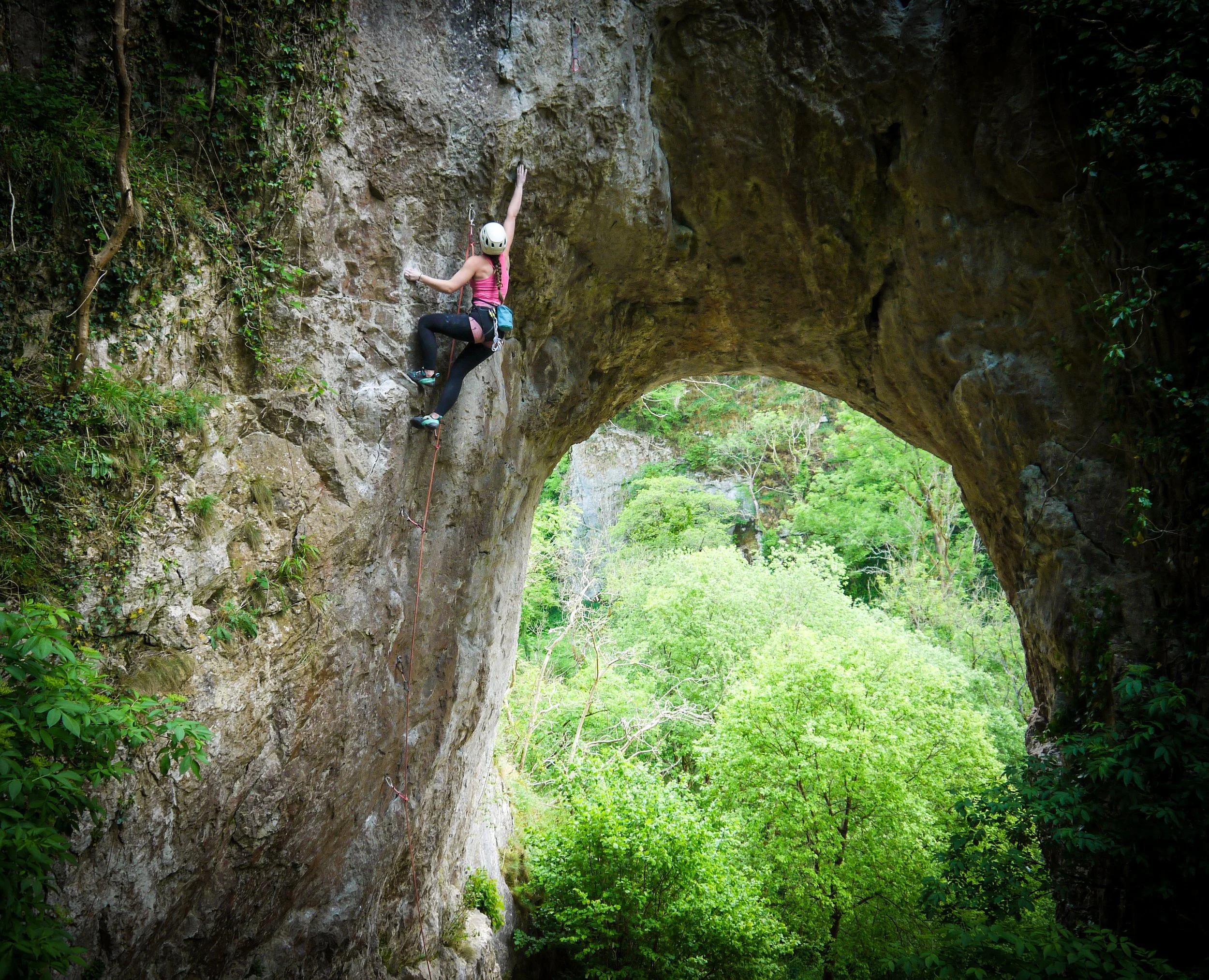 A woman rock climbing on a vertical rock face with a green forest in the background, wearing a white helmet, pink tank top, black leggings, and climbing shoes.