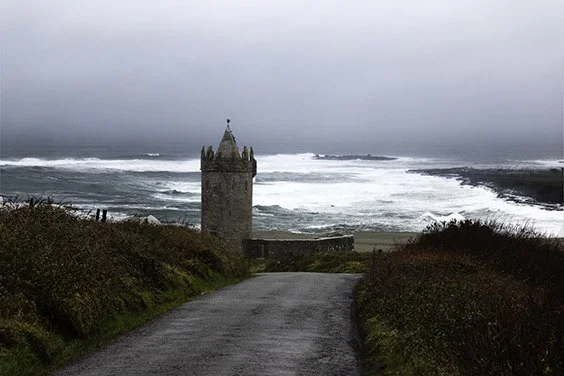A narrow road leading to Doonagore a 16th-century tower house with a small walled enclosure located on the Clare coast near Doolin, with turbulent ocean waves and cloudy sky in the background.