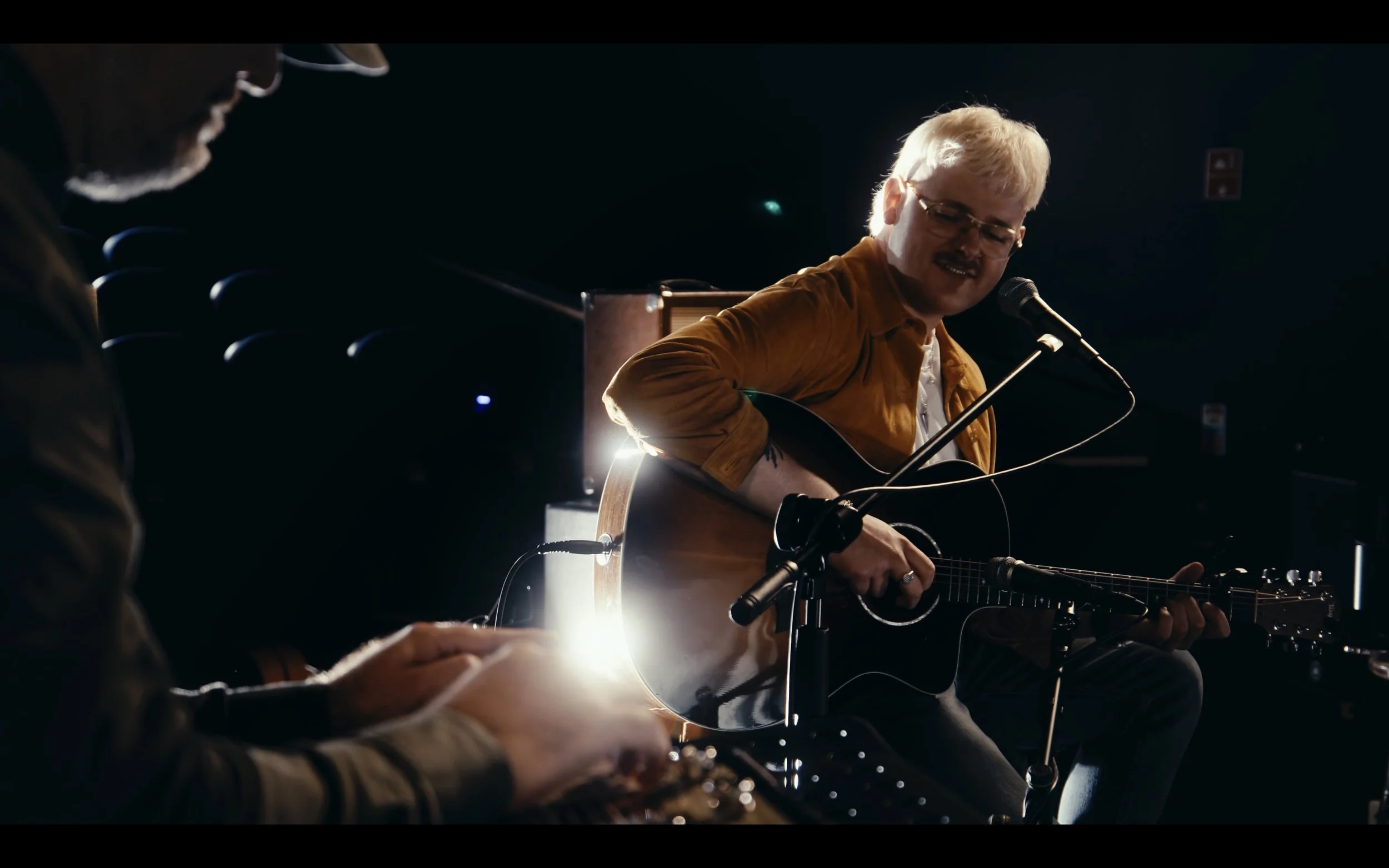 A man with blond hair and glasses playing an acoustic guitar and singing into a microphone while a person on the left plays a keyboard. The scene is dimly lit with backlighting highlighting the performers.
