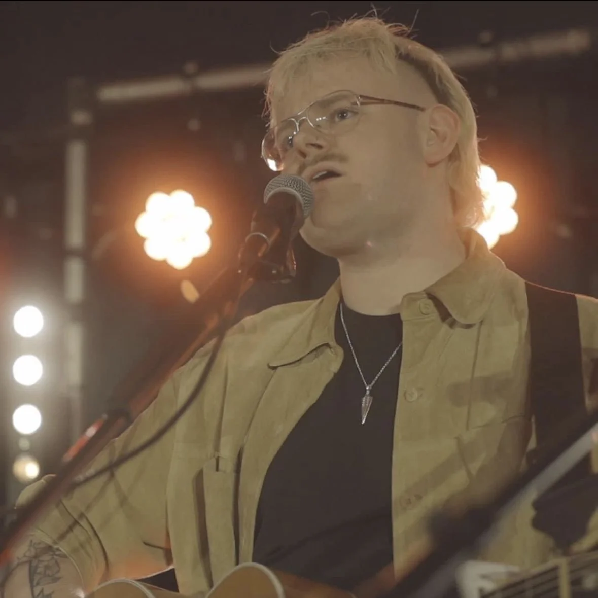 A man singing into a microphone while playing an acoustic guitar, with stage lights in the background.