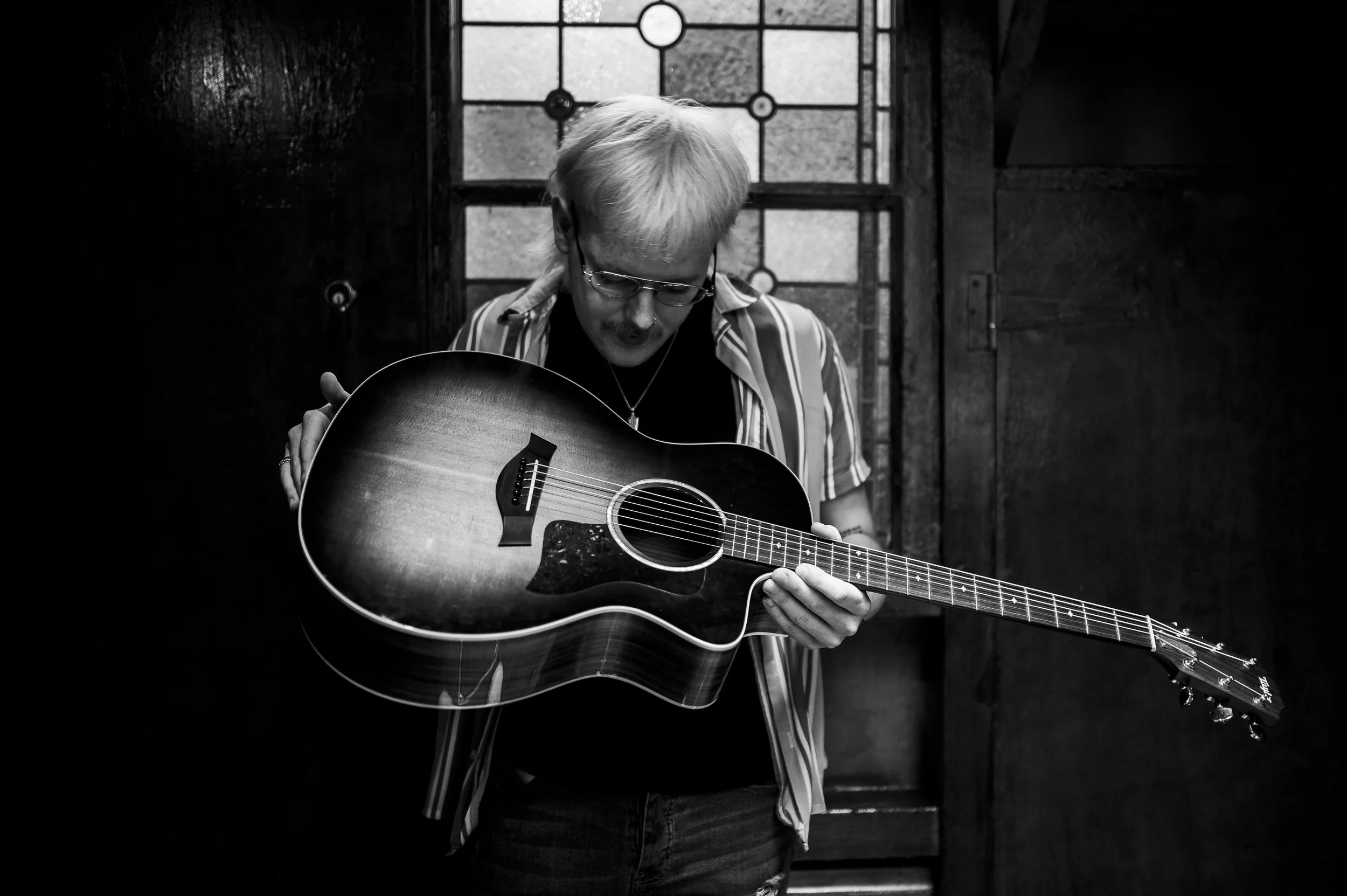 A person with light-colored hair, glasses, and a striped shirt holding an acoustic guitar, standing in front of a stained glass window in a dimly lit room.