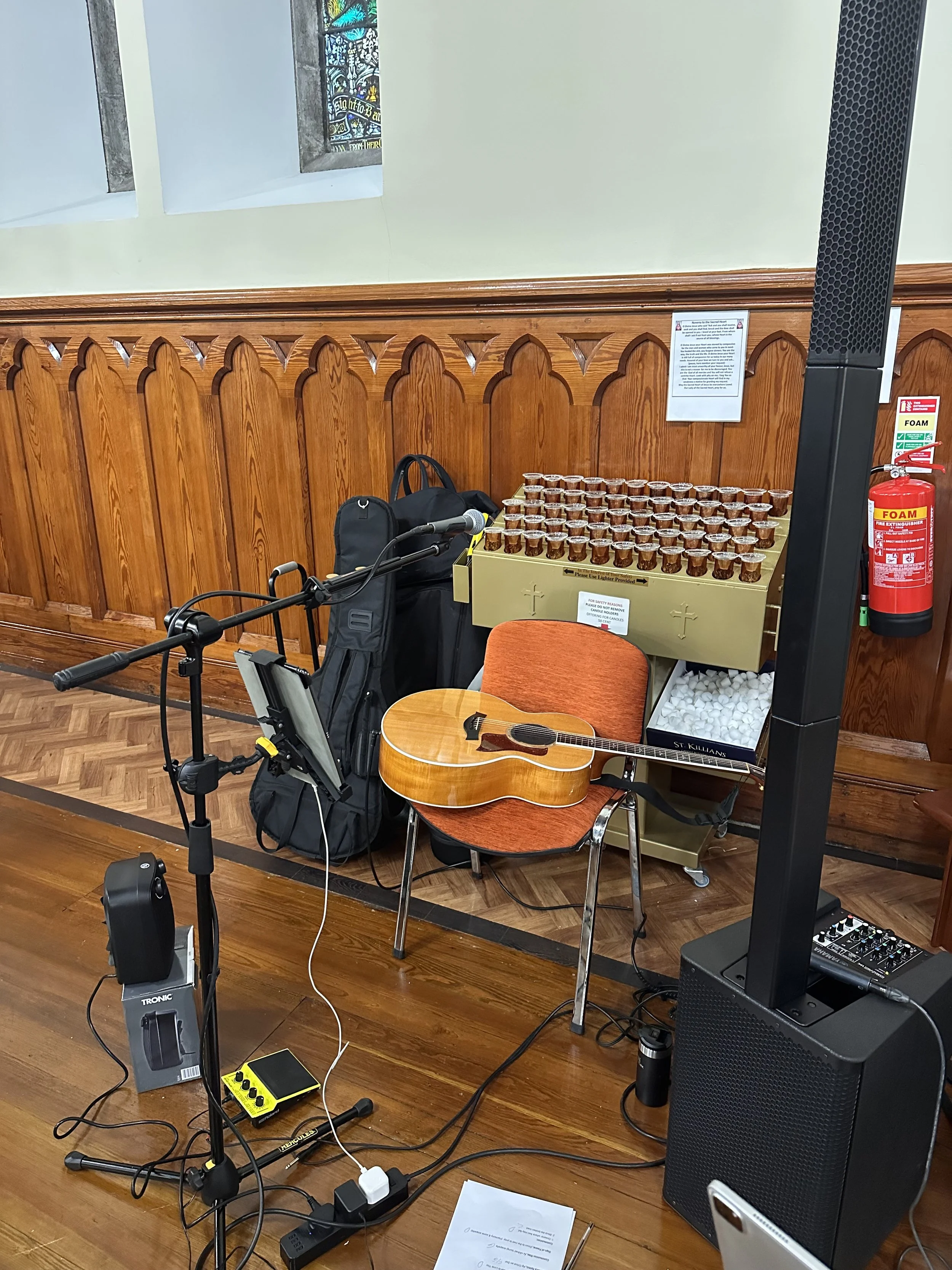 Musician setup with an acoustic guitar on a chair, microphone and sound equipment, a backpack, a box of small cups, and a fire extinguisher inside a room with wooden paneling and stained glass windows.