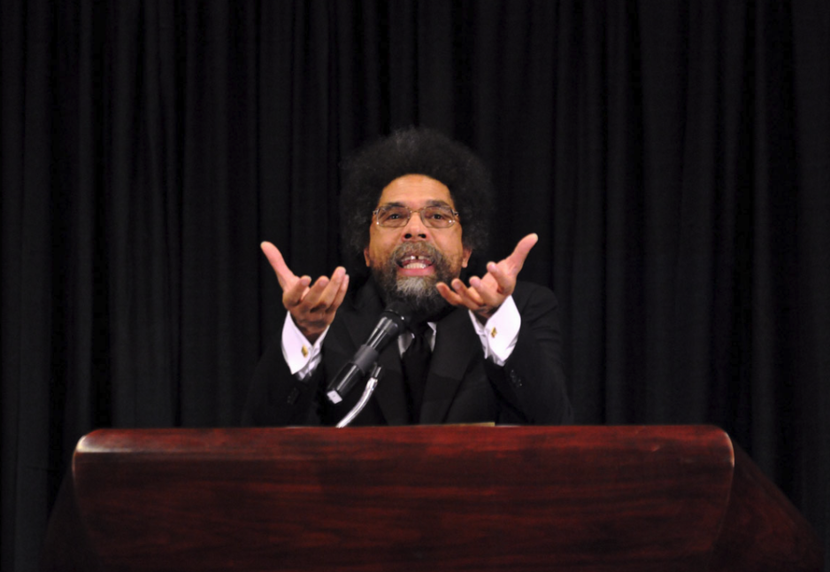 Dr. Cornel West speaking at a podium with a black curtain in the background, gesturing with his hands.