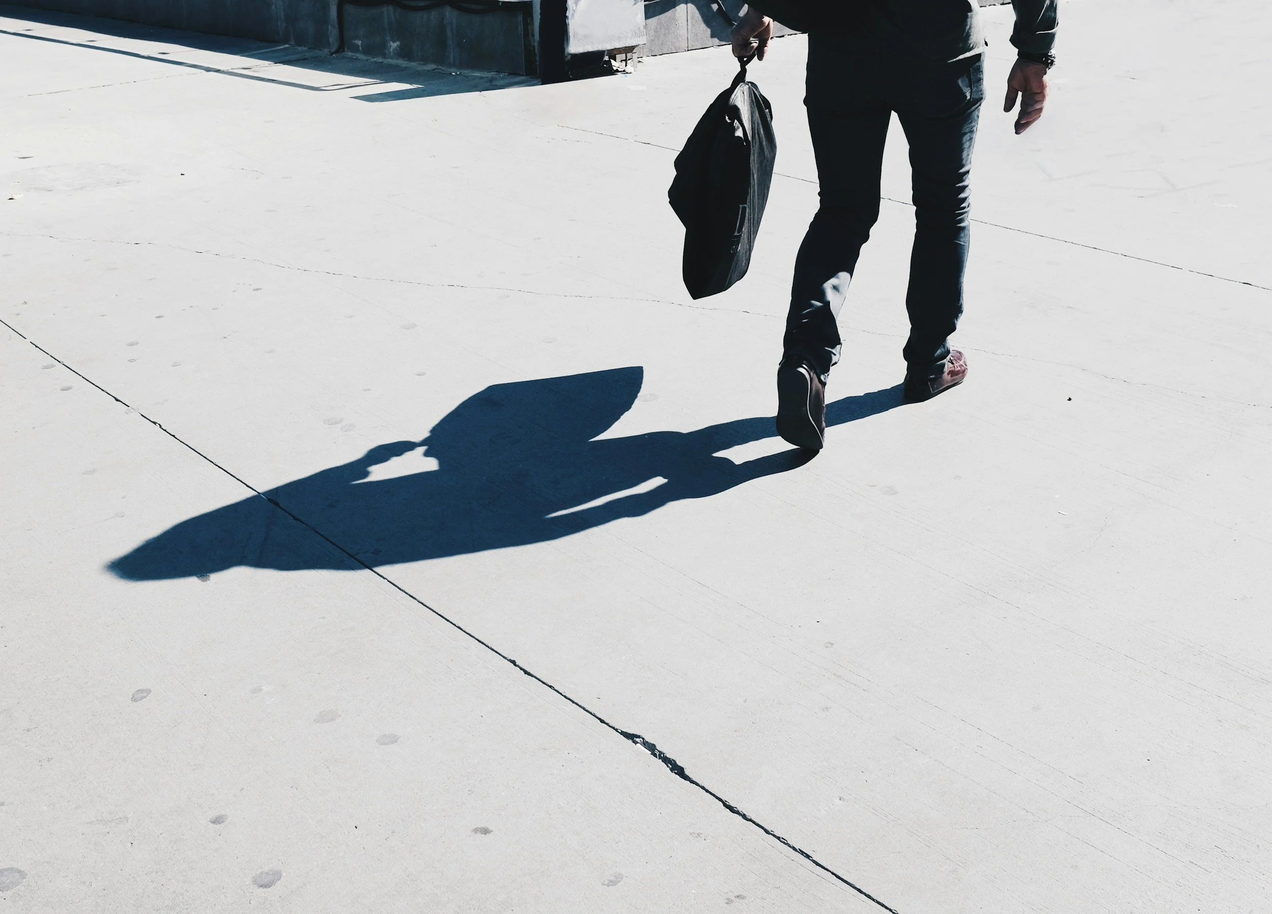 A person walking on a sidewalk carrying a shoulder bag, with their shadow cast onto the concrete.