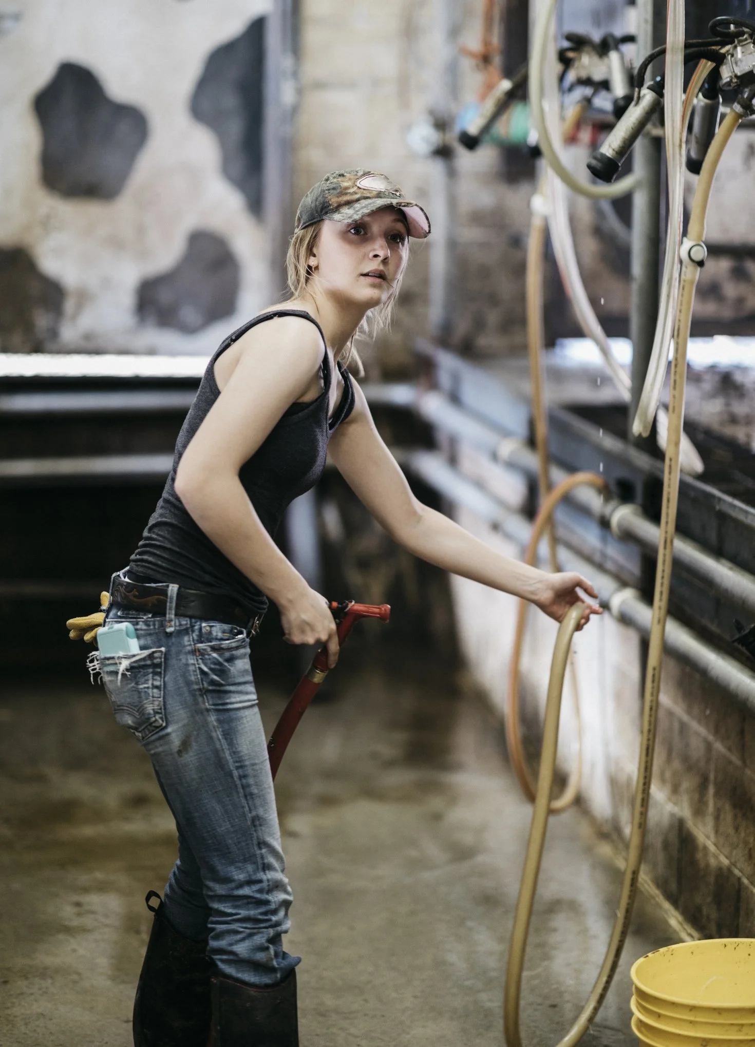A young woman wearing a camouflage baseball cap, a sleeveless black top, and jeans backed with tools, is washing or cleaning in a barn or farm setting. She is holding a tool and standing near a wall with pipes and hoses.