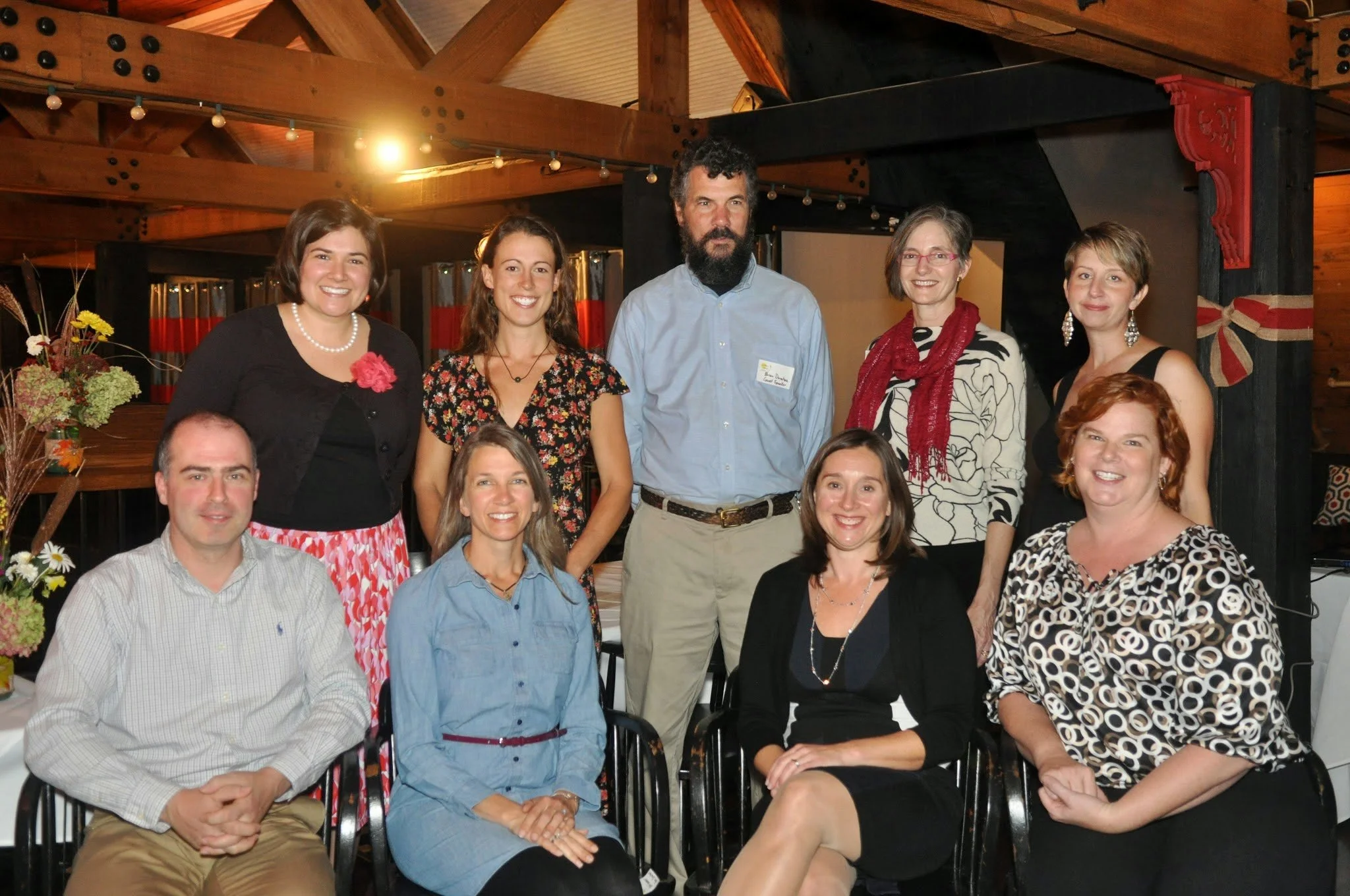 A group of ten people, five women and five men, posing together in a rustic, wooden indoor setting with string lights. They are dressed in casual and semi-formal attire, smiling at the camera.