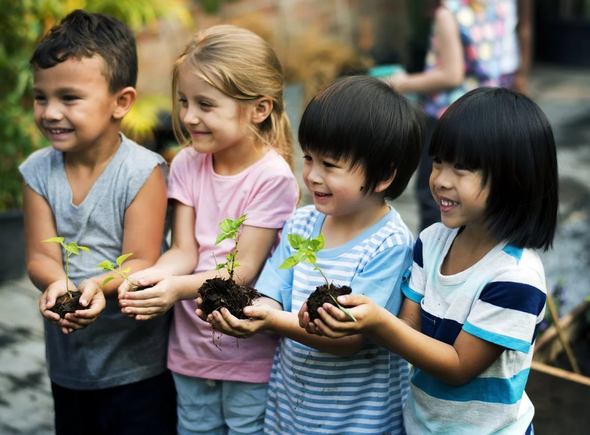 Four children holding small potted plants outdoors, smiling.