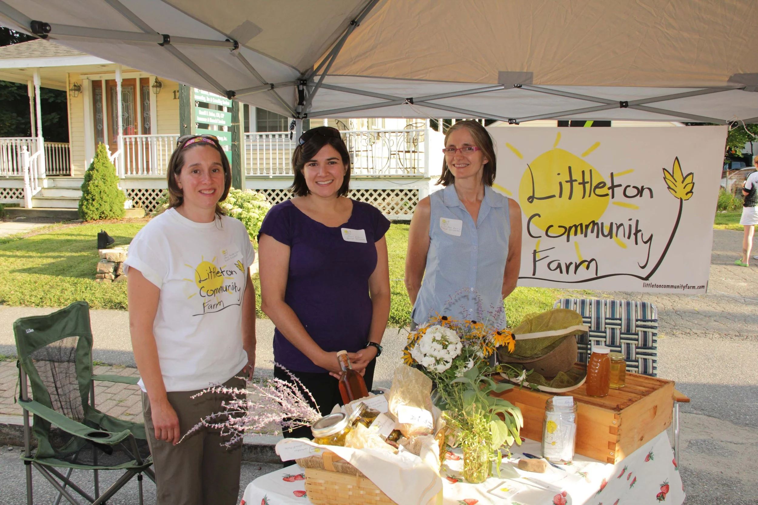 Three women behind a table at the Littleton Community Farm booth during an outdoor event, with a banner displaying the farm's name. The table has jars of honey, flowers, and other farm products. The women are smiling and standing under a canopy with a residential neighborhood in the background.